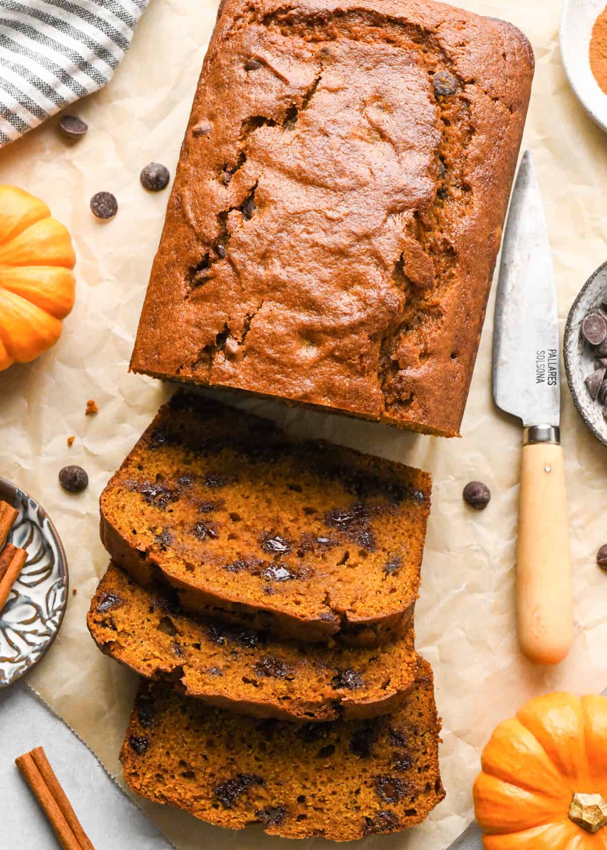 a loaf of pumpkin chocolate chip bread with 3 slices cut out of it on parchment paper, with melty chocolate chips surrounded by mini pumpkins, cinnamon sticks, a knife and a bowl of chocolate chips