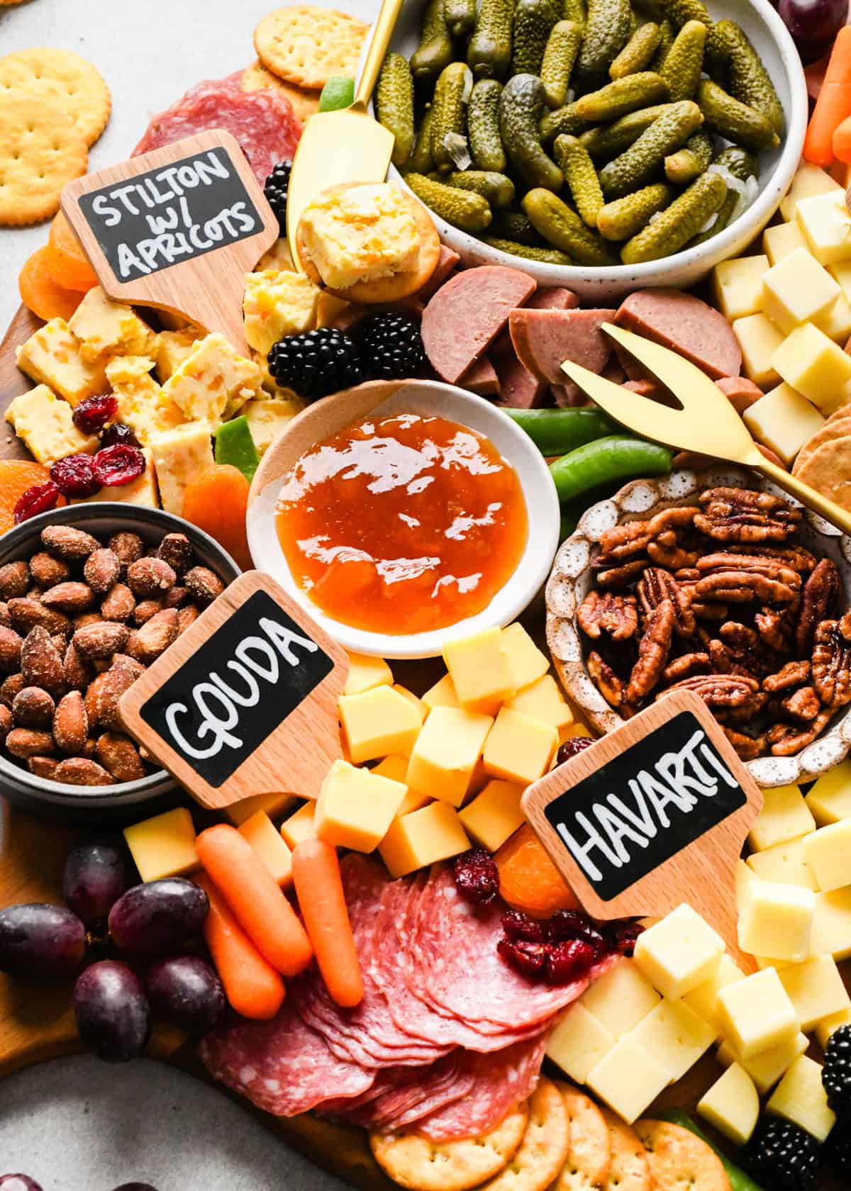overhead photo of a charcuterie board with assortment of cubed cheeses with chalkboard labels reading Gouda, Havarti and Stilton with apricots), green olives, pub cheese spread, salami, summer sausage, pickles, nuts, dried and fresh fruits, crackers, and a bowl of apricot jam, all arranged on a round wooden serving board.