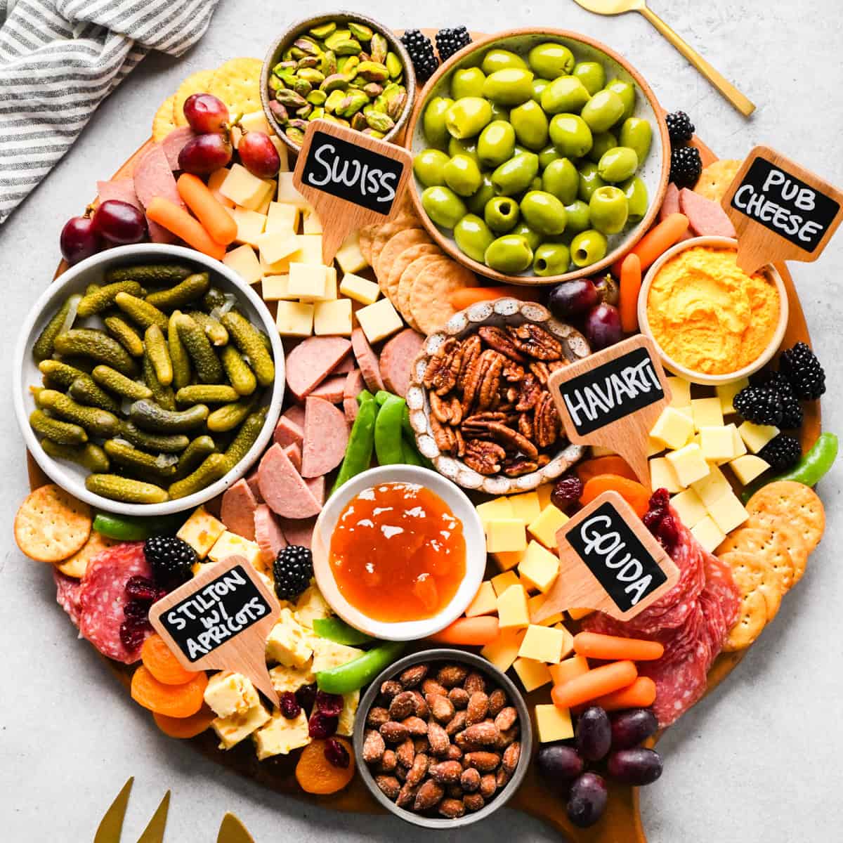 overhead photo of a charcuterie board with assortment of cubed cheeses with chalkboard labels reading Swiss, Gouda, Havarti and Stilton with apricots), green olives, pub cheese spread, salami, summer sausage, pickles, nuts, dried and fresh fruits, crackers, and a bowl of apricot jam, all arranged on a round wooden serving board.