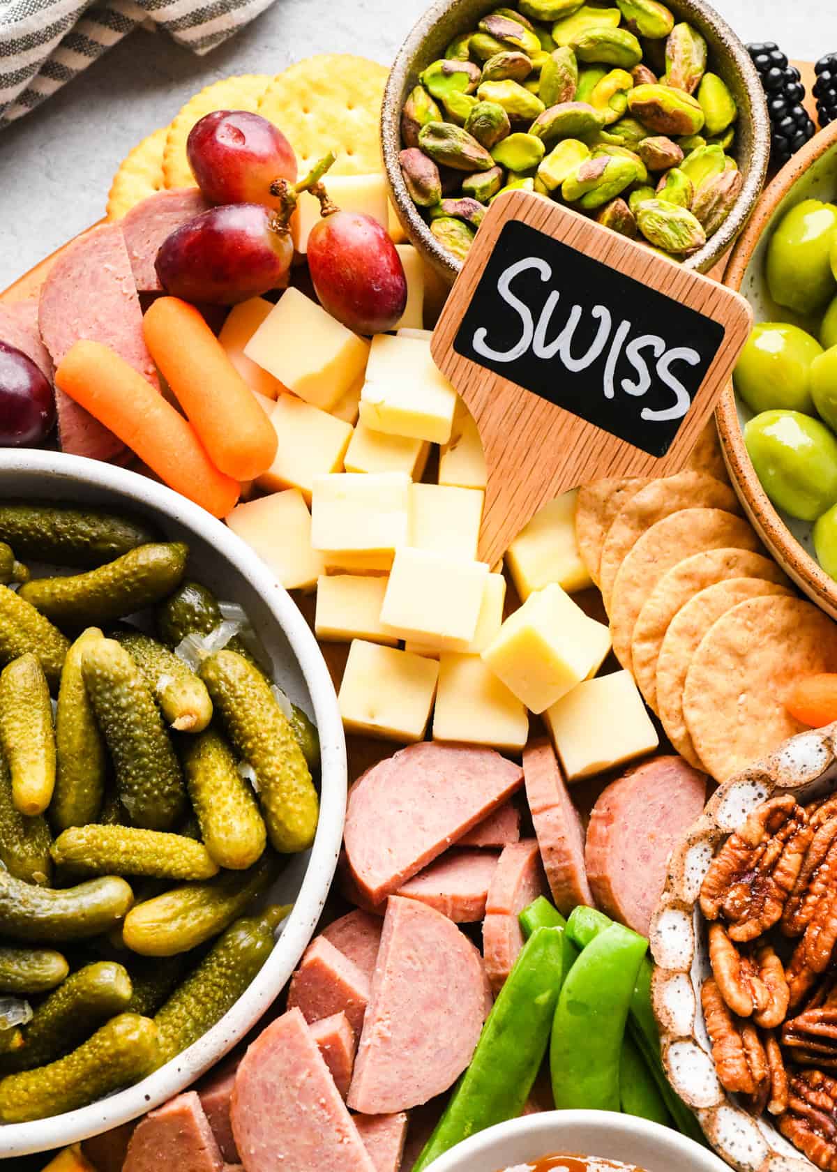 up close photo of a portion of a charcuterie board with a cheese marker labeled "swiss," pickles, pistachios, grapes, carrots, summer sausage, green olives and pecans