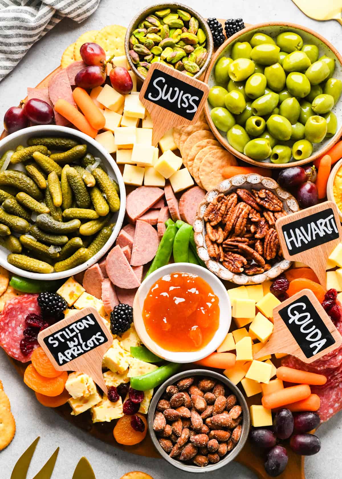 up close overhead photo of a charcuterie board with assortment of cubed cheeses with chalkboard labels reading Swiss, Gouda, Havarti and Stilton with apricots), green olives, pub cheese spread, salami, summer sausage, pickles, nuts, dried and fresh fruits, crackers, and a bowl of apricot jam, all arranged on a round wooden serving board.