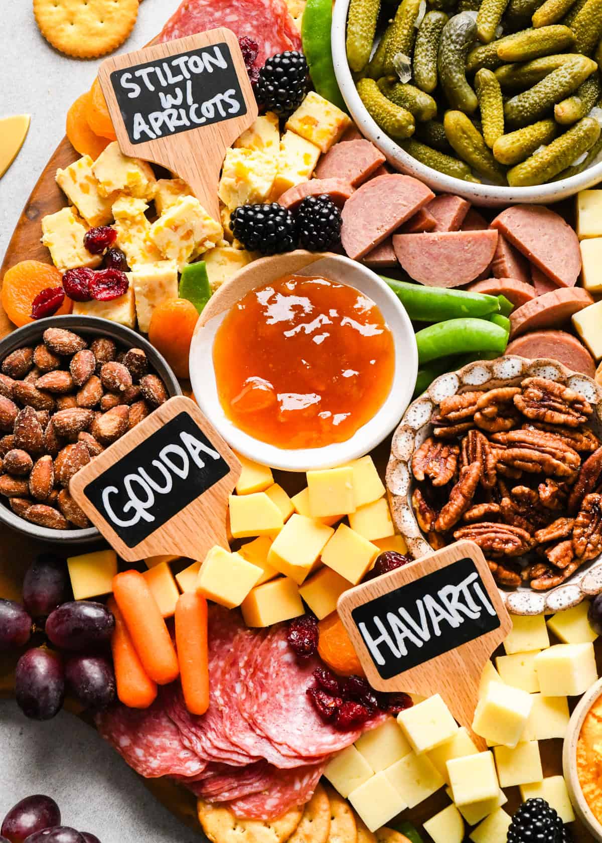 overhead photo of a charcuterie board with assortment of cubed cheeses with chalkboard labels reading Swiss, Gouda, Havarti and Stilton with apricots), green olives, pub cheese spread, salami, summer sausage, pickles, nuts, dried and fresh fruits, crackers, and a bowl of apricot jam, all arranged on a round wooden serving board.