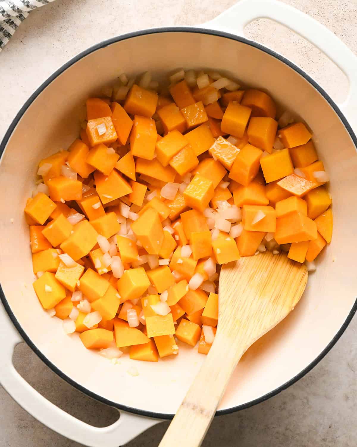 Making butternut squash risotto - butternut squash, garlic, onion, and butter in a white 5-quart saucepan being stirred with a wooden spoon