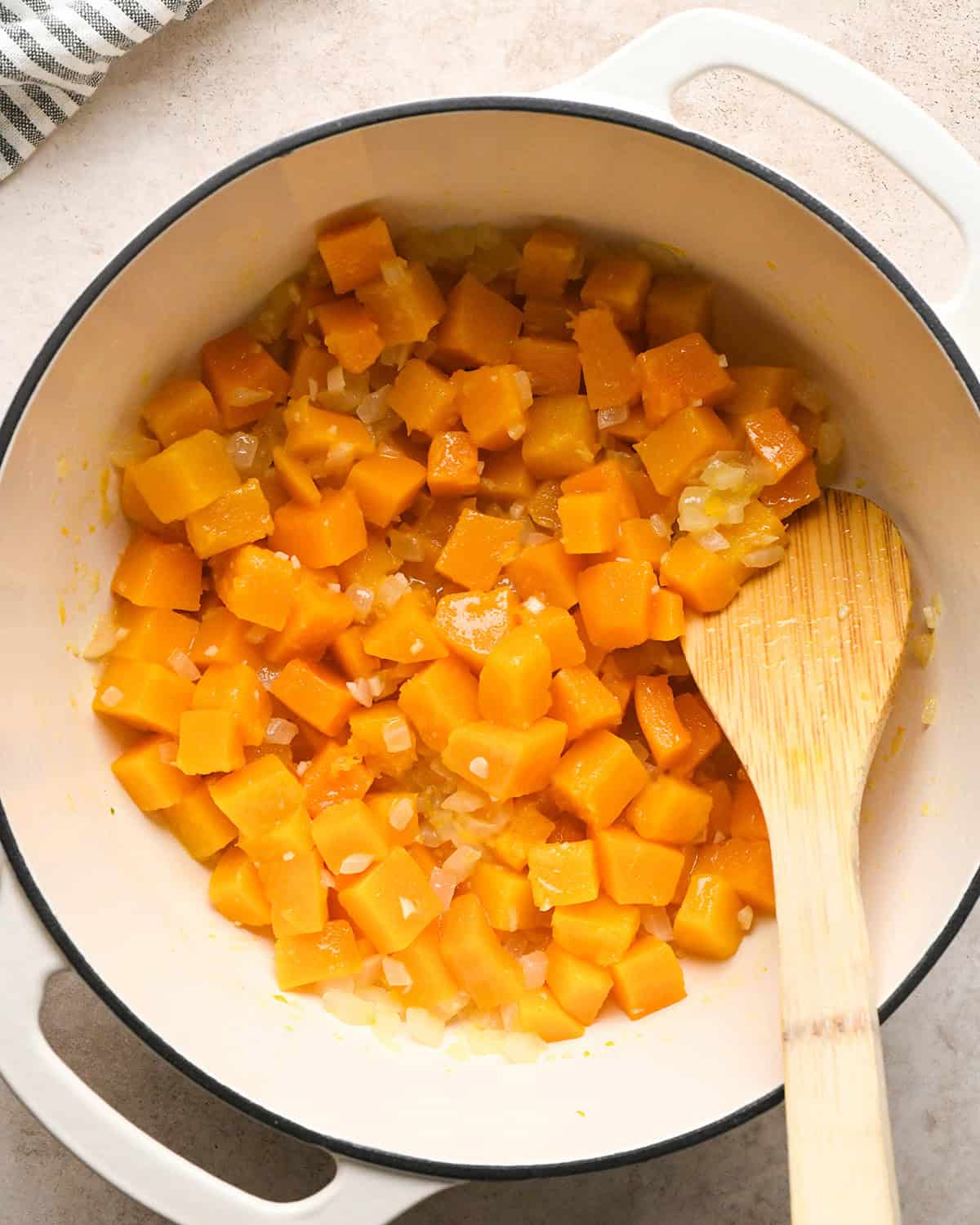 Making butternut squash risotto - butternut squash, garlic, onion, and butter in a white 5-quart saucepan being stirred with a wooden spoon