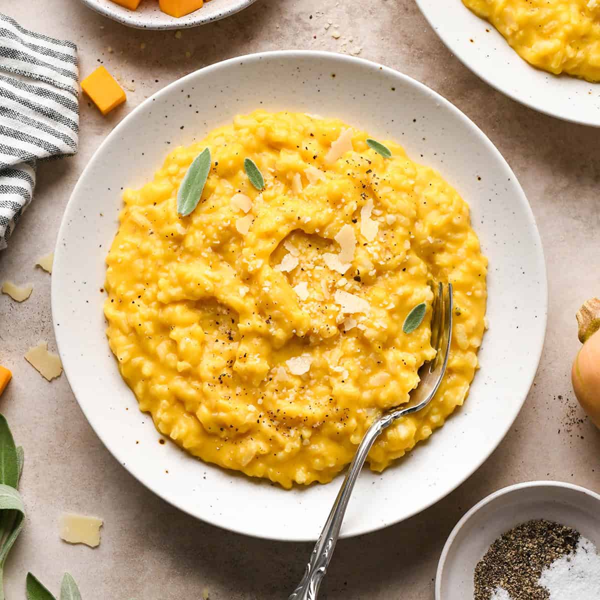 butternut squash risotto garnished with shaved parmesan cheese, sage leaves, and black pepper on a white speckled plate with a fork surrounded by parmesan cheese, butternut squash and another bowl of risotto