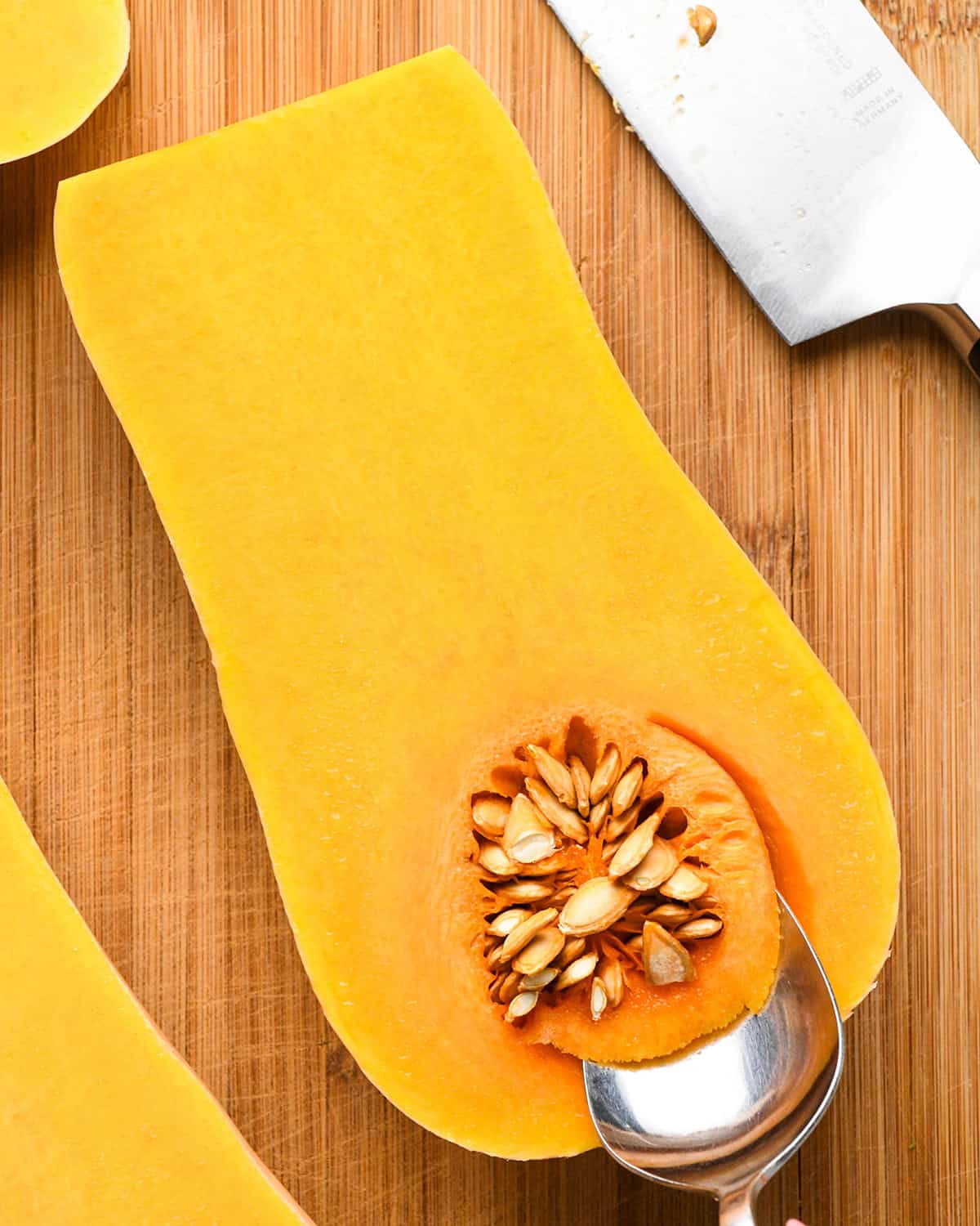 Halved butternut squash with seeds being scooped out with a spoon - on a wooden cutting board next to a knife to be used to make butternut squash risotto