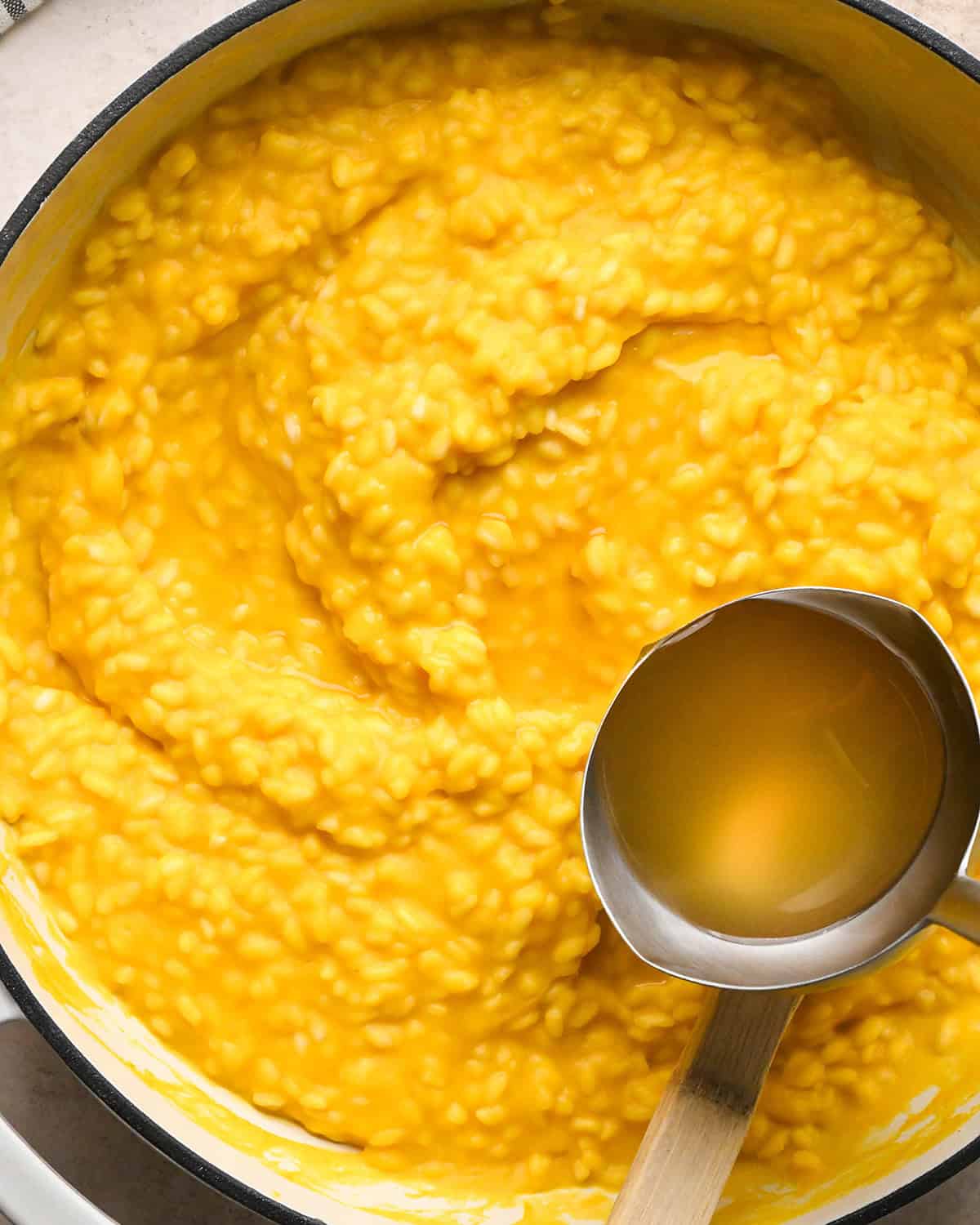Making butternut squash risotto - all ingredients in a white saucepan, adding chicken broth with a silver ladle 