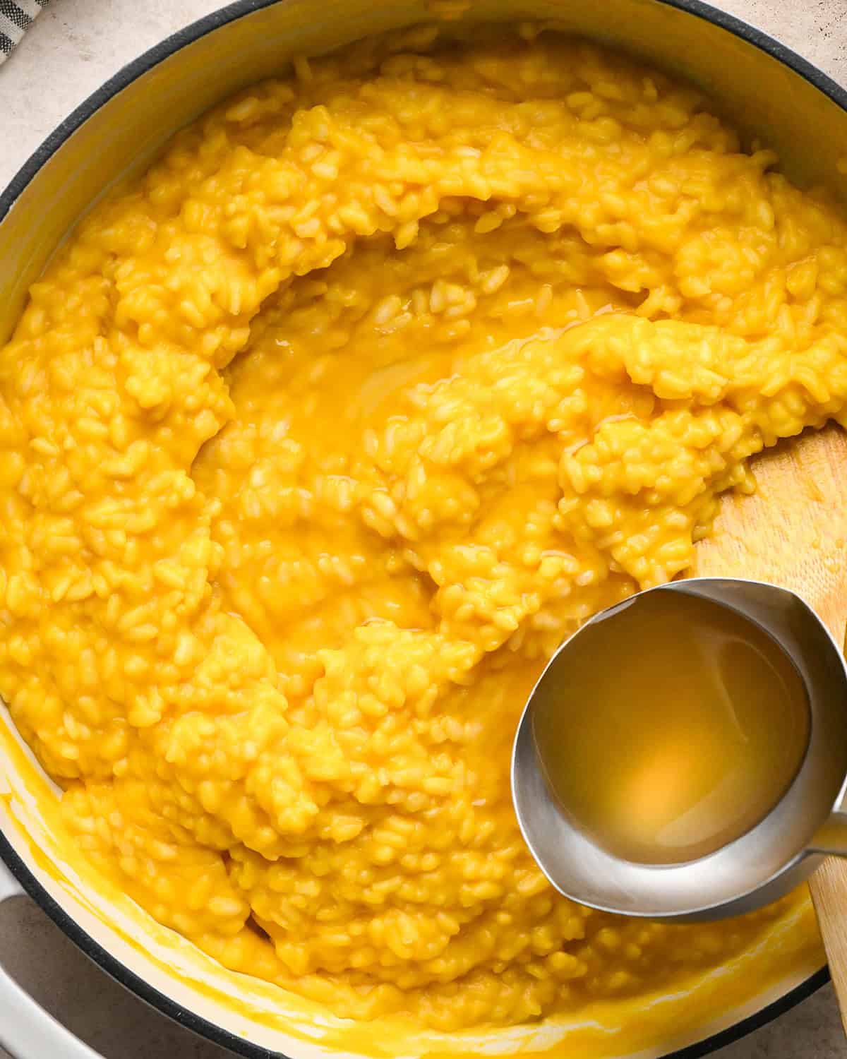Making butternut squash risotto - all ingredients in a white saucepan, adding chicken broth with a silver ladle 