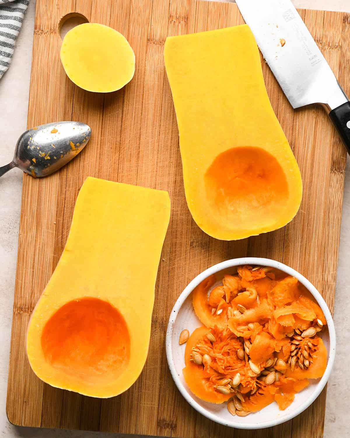 Halved butternut squash with seeds scooped out and placed in a bowl - on a wooden cutting board next to a knife and a spoon to be used to make butternut squash risotto
