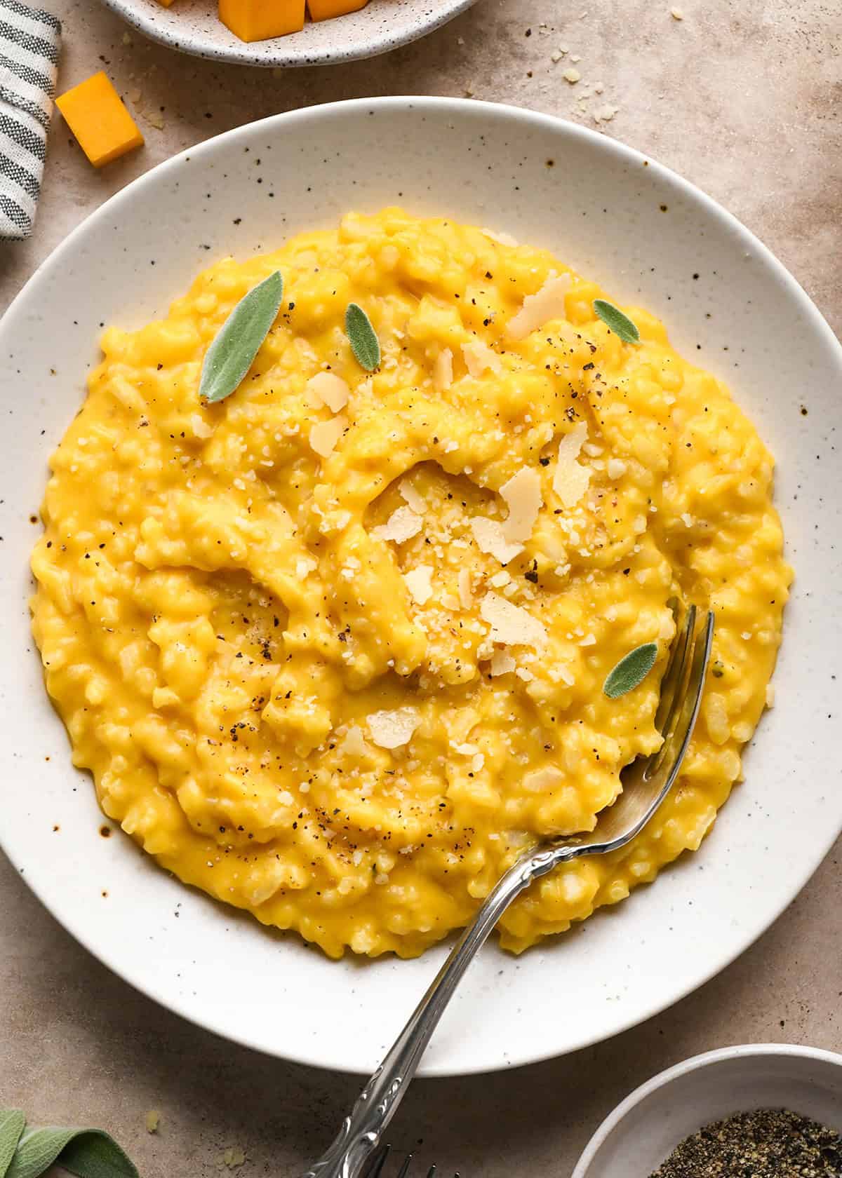 overhead photo of butternut squash risotto served on a white speckled plate with a fork and topped with sage, parmesan cheese, and pepper