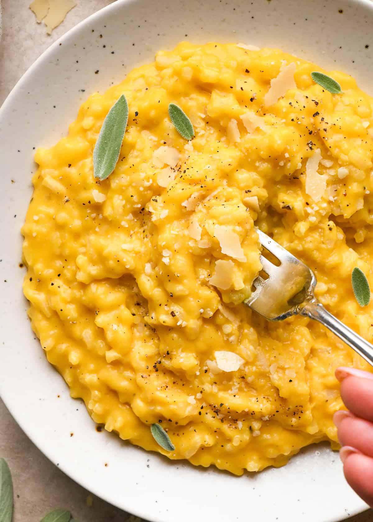 overhead photo of butternut squash risotto served on a white speckled plate with a fork and topped with sage, parmesan cheese, and pepper