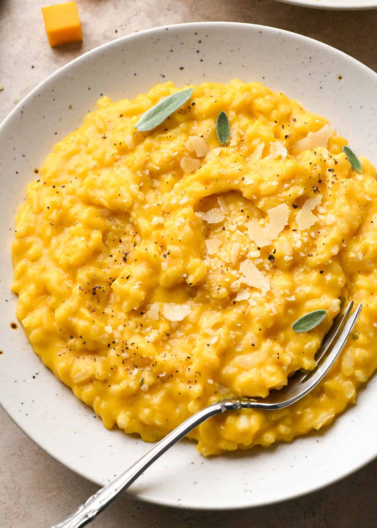 Creamy butternut squash risotto topped with shaved parmesan, cracked pepper, and sage on a white speckled plate with a fork