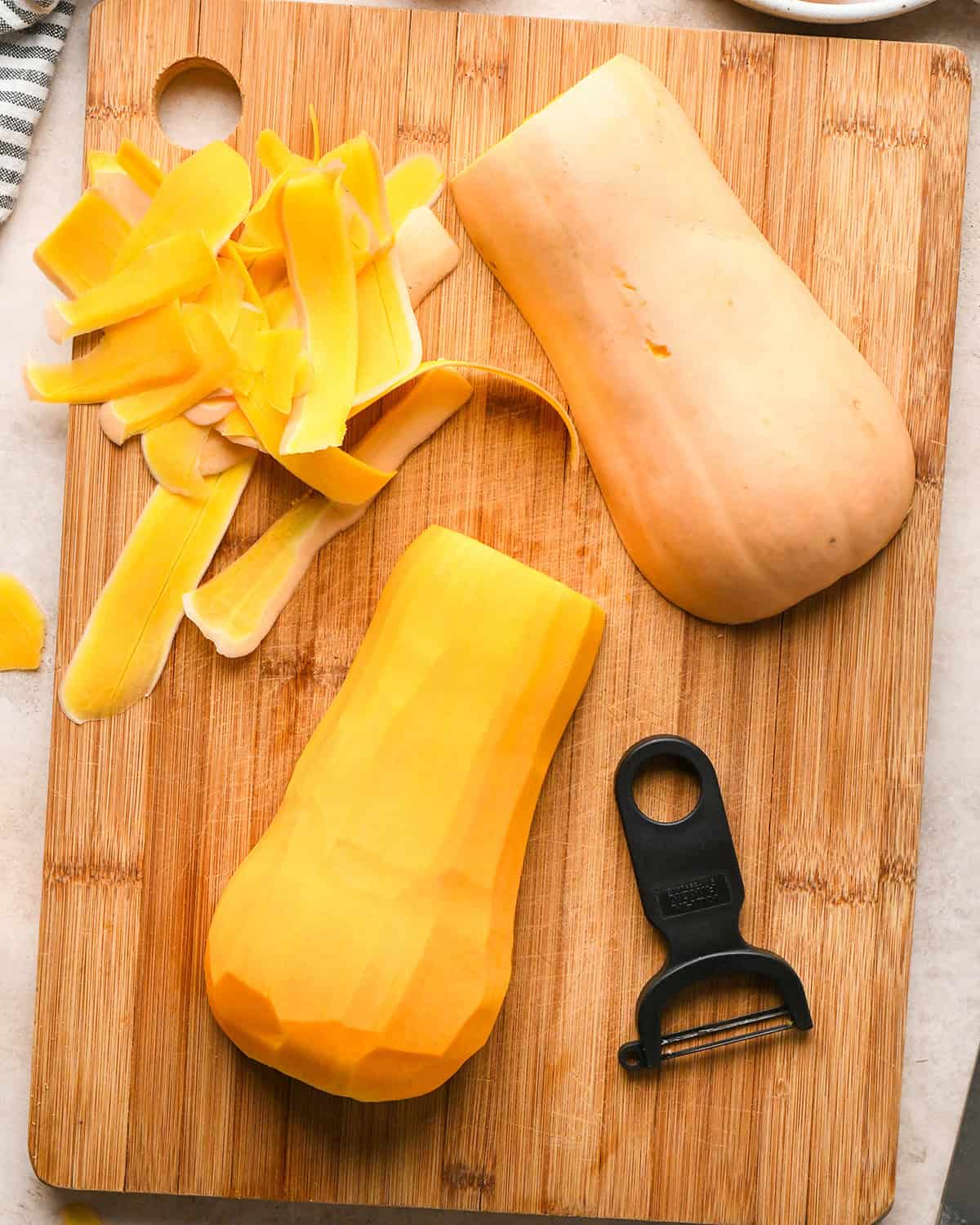 Peeled butternut squash on a wooden cutting board next to a vegetable peeler to be used to make butternut squash risotto