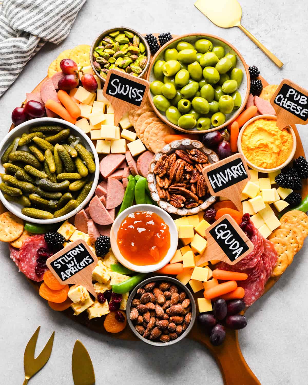 overhead photo of a charcuterie board with assortment of cubed cheeses with chalkboard labels reading Swiss, Gouda, Havarti and Stilton with apricots), green olives, pub cheese spread, salami, summer sausage, pickles, nuts, dried and fresh fruits, crackers, and a bowl of apricot jam, all arranged on a round wooden serving board.