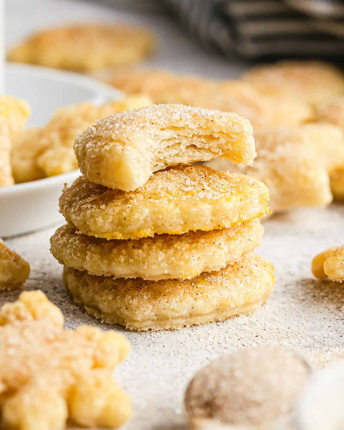 a stack of 4 Pie Crust Cookies coated in cinnamon sugar, the top one has a bite taken out of it, surrounded by other cookies