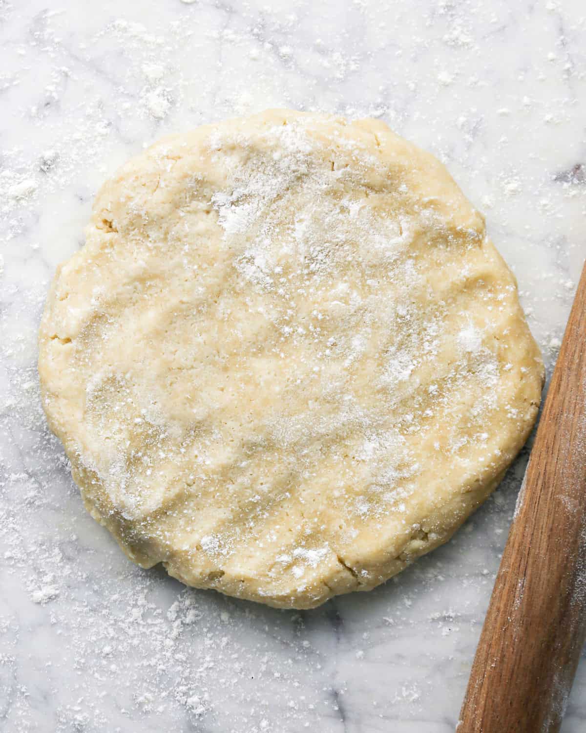 a disc of pie crust dough on a gray, marble surface sprinkled with flour with a wooden rolling pin next to it to make pie crust cookies