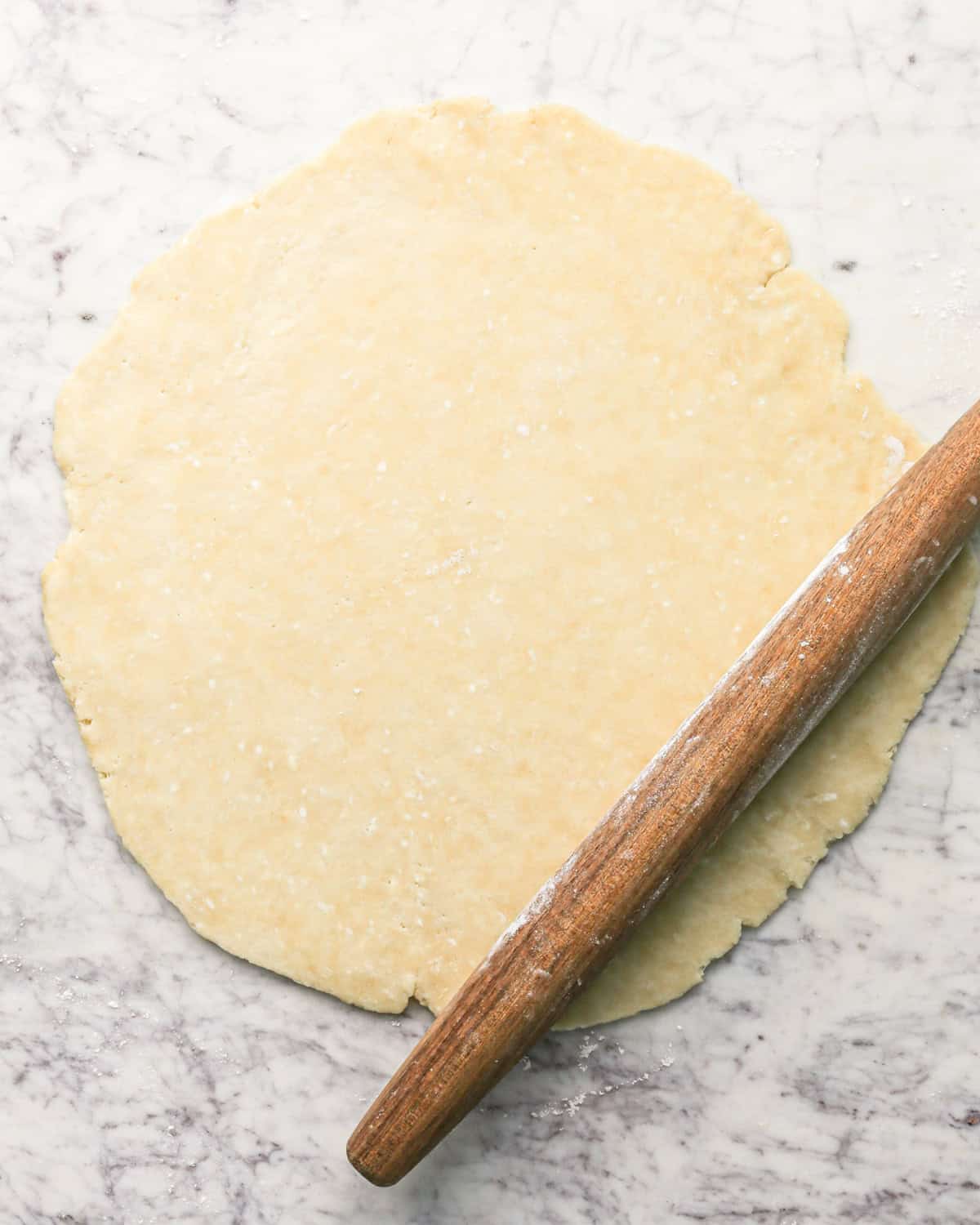 pie crust dough rolled into a thin circle on a gray, marble surface sprinkled with flour with a wooden rolling pin next to it to make pie crust cookies