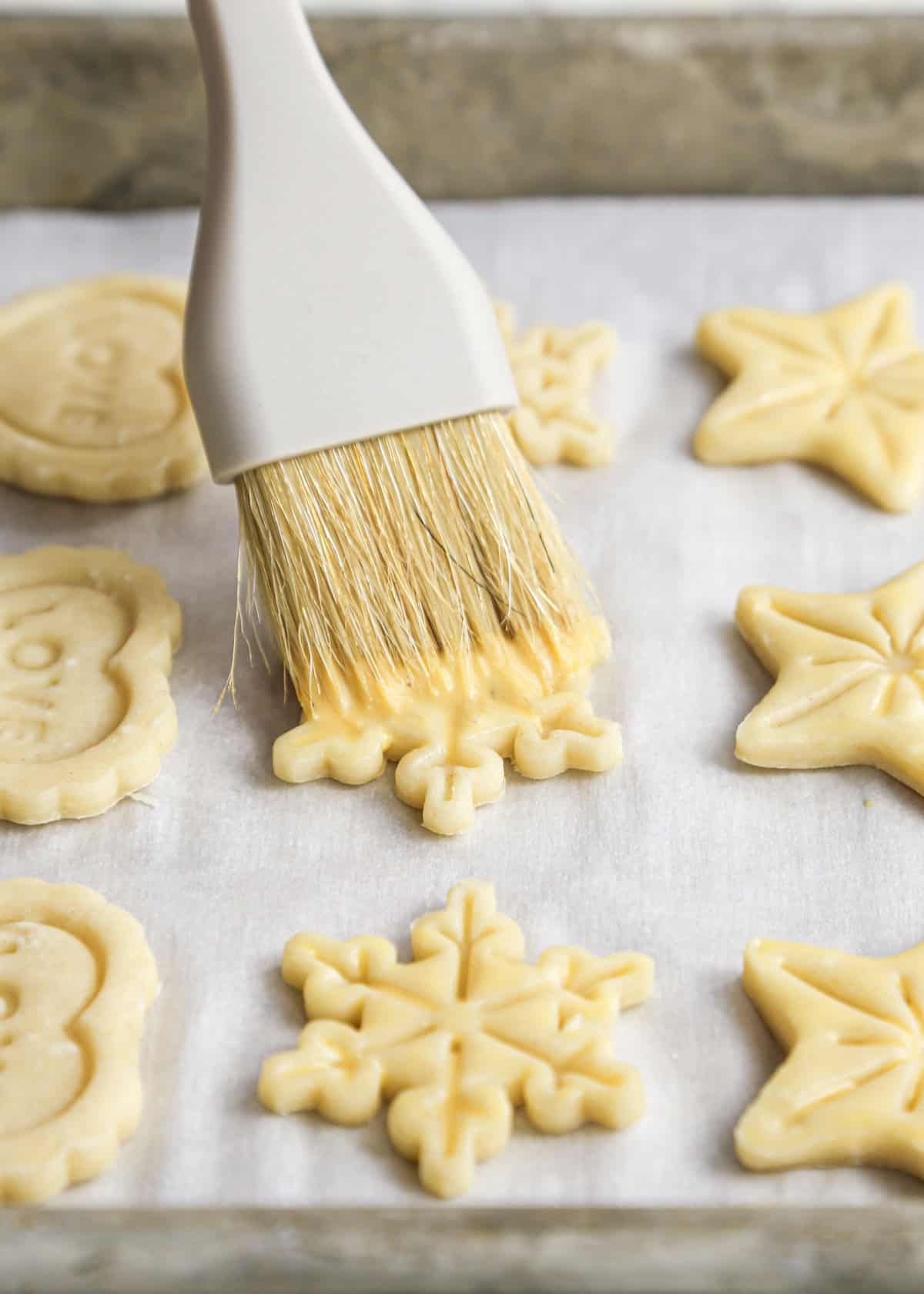 a pastry brush brushing egg wash on 3 rows of pie crust cookies cut into stars, snowflakes and hearts on a baking sheet lined with parchment paper before baking