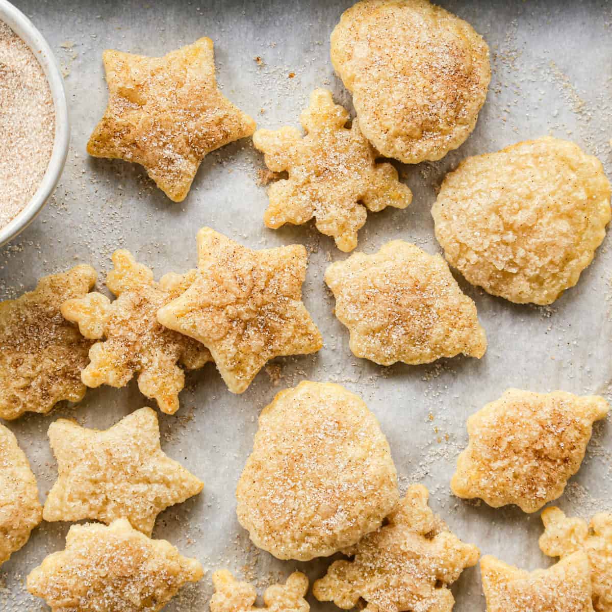 15 Pie Crust Cookies with cinnamon sugar in the shapes of hearts, stars, leaves and snowflakes on a gray surface with a bowl of cinnamon sugar in the top left corner