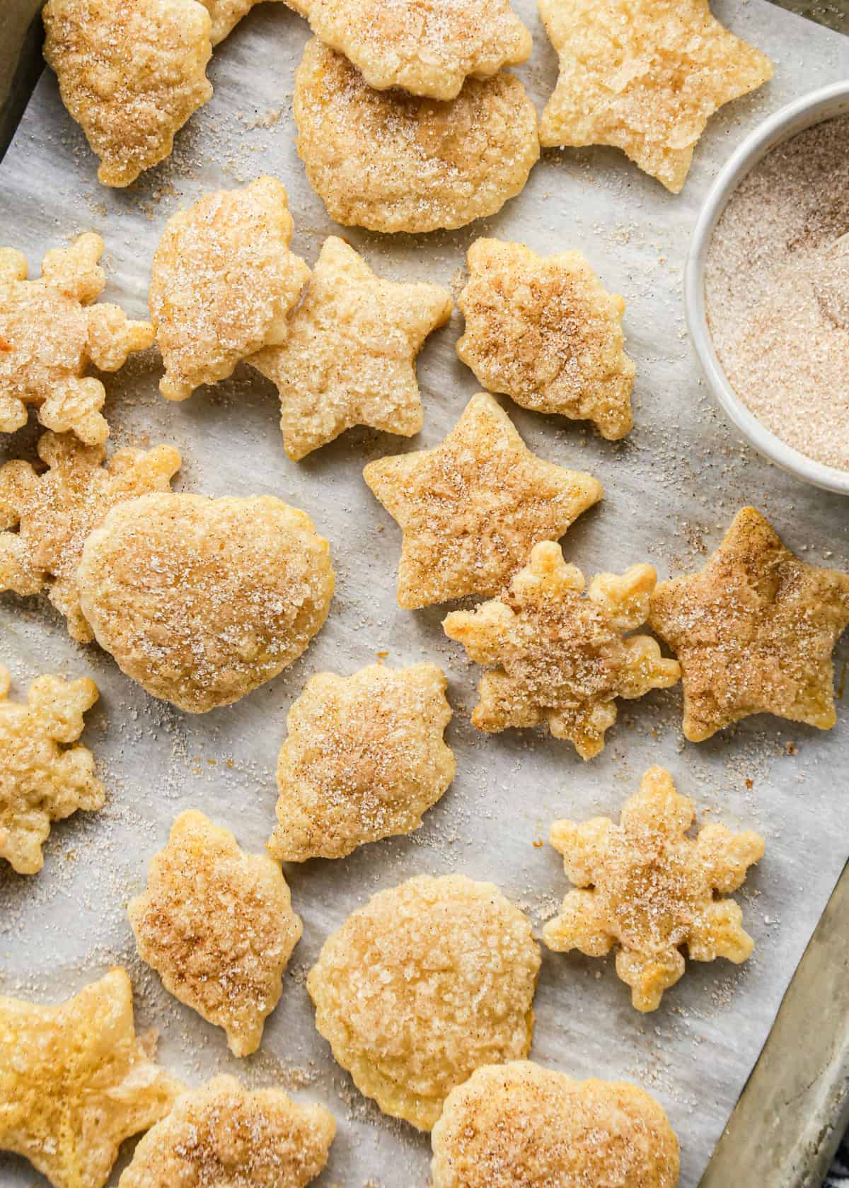 21 Pie Crust Cookies with cinnamon sugar in the shapes of hearts, stars, leaves and snowflakes on a parchment paper lined baking sheet with a bowl of cinnamon sugar in the top right corner