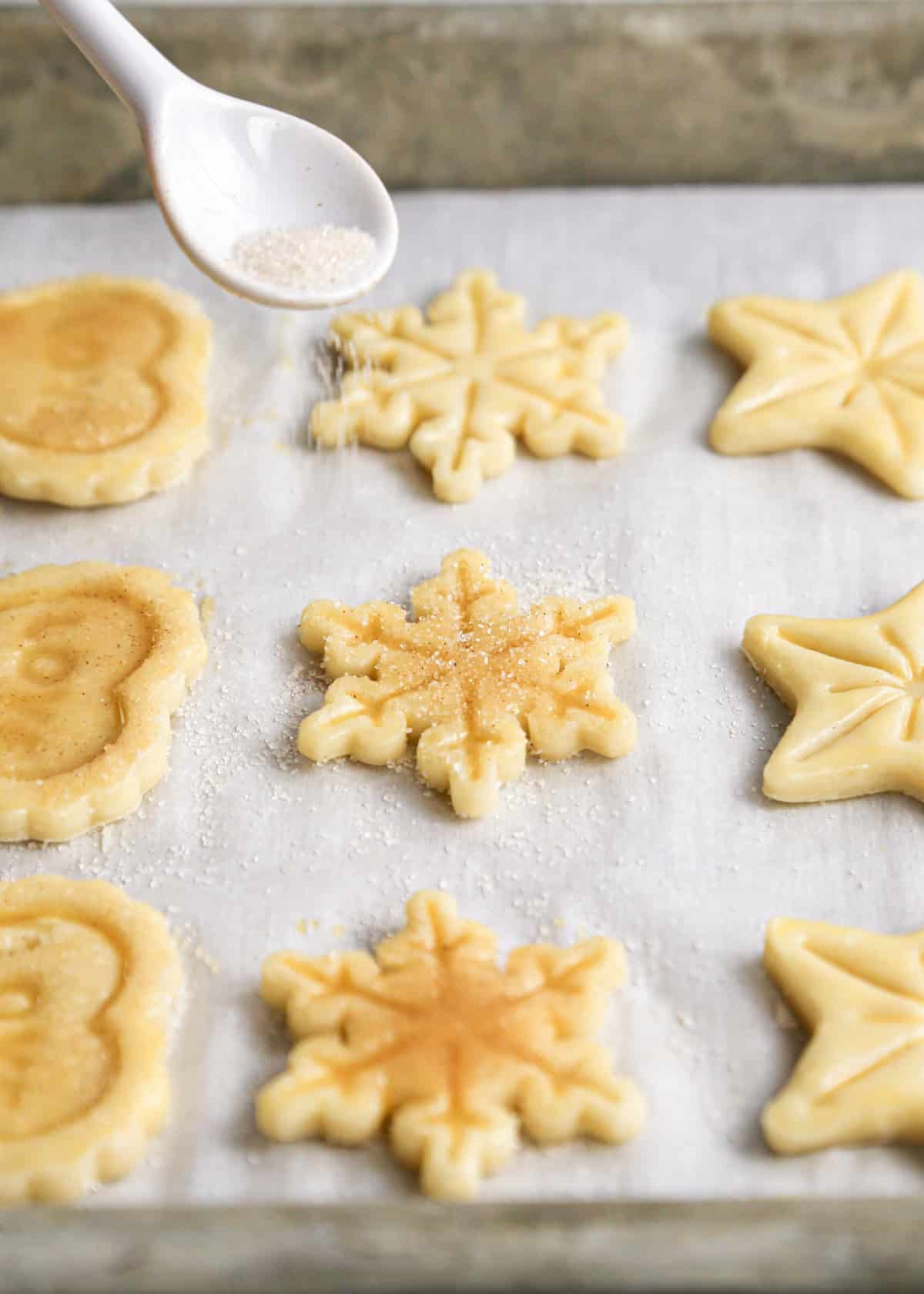 a spoon sprinkling cinnamon sugar over 3 rows of pie crust cookies cut into stars, snowflakes and hearts on a baking sheet lined with parchment paper before baking