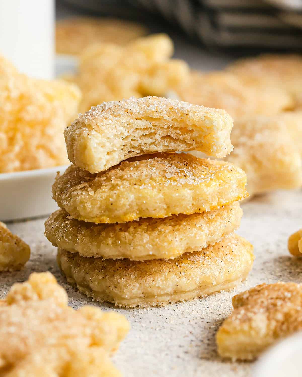 a stack of 4 Pie Crust Cookies coated in cinnamon sugar, the top one has a bite taken out of it, surrounded by other cookies