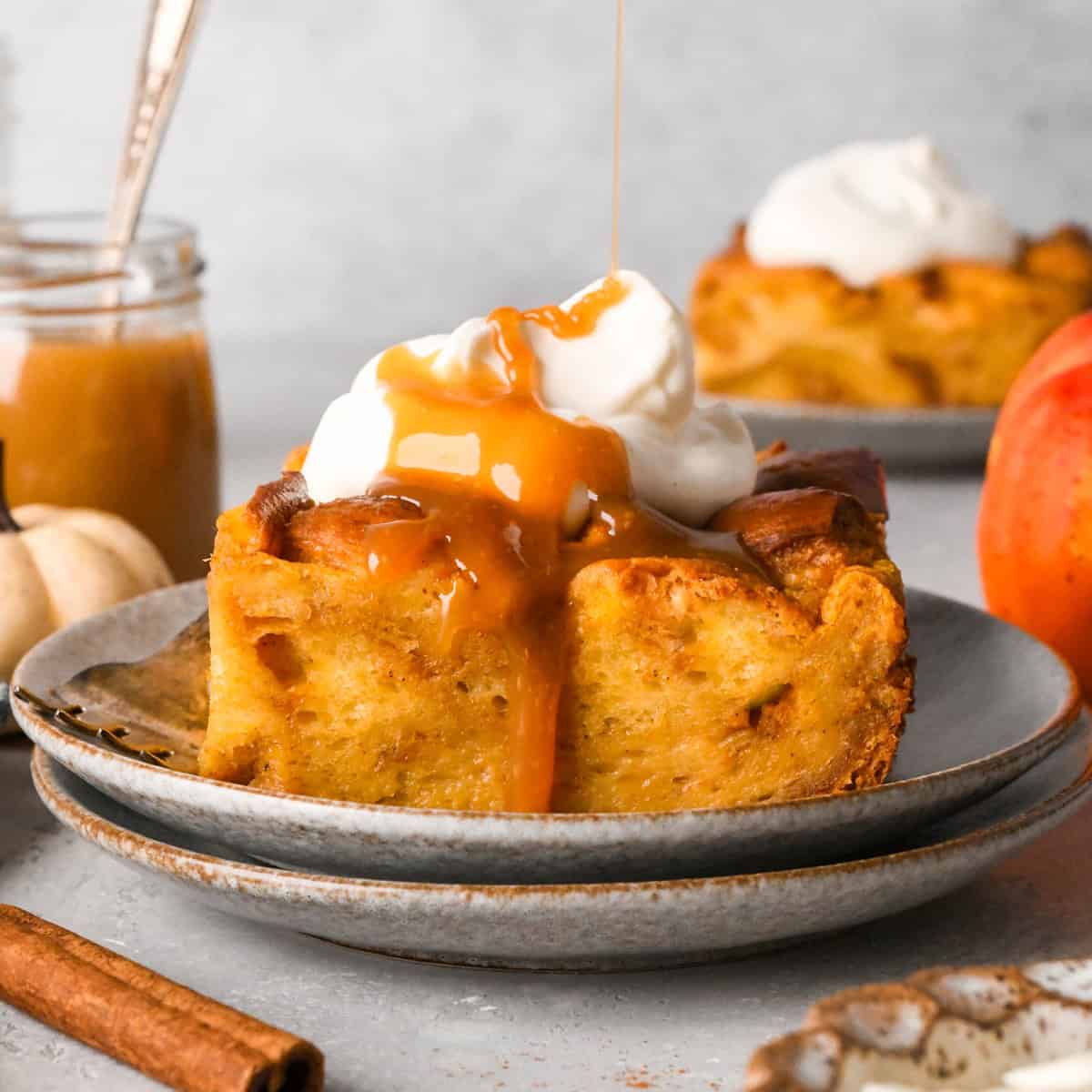a piece of Pumpkin Bread Pudding on a plate with whipped cream and caramel sauce with another piece on a plate in the background, surrounded by caramel and pumpkins and cinnamon sticks