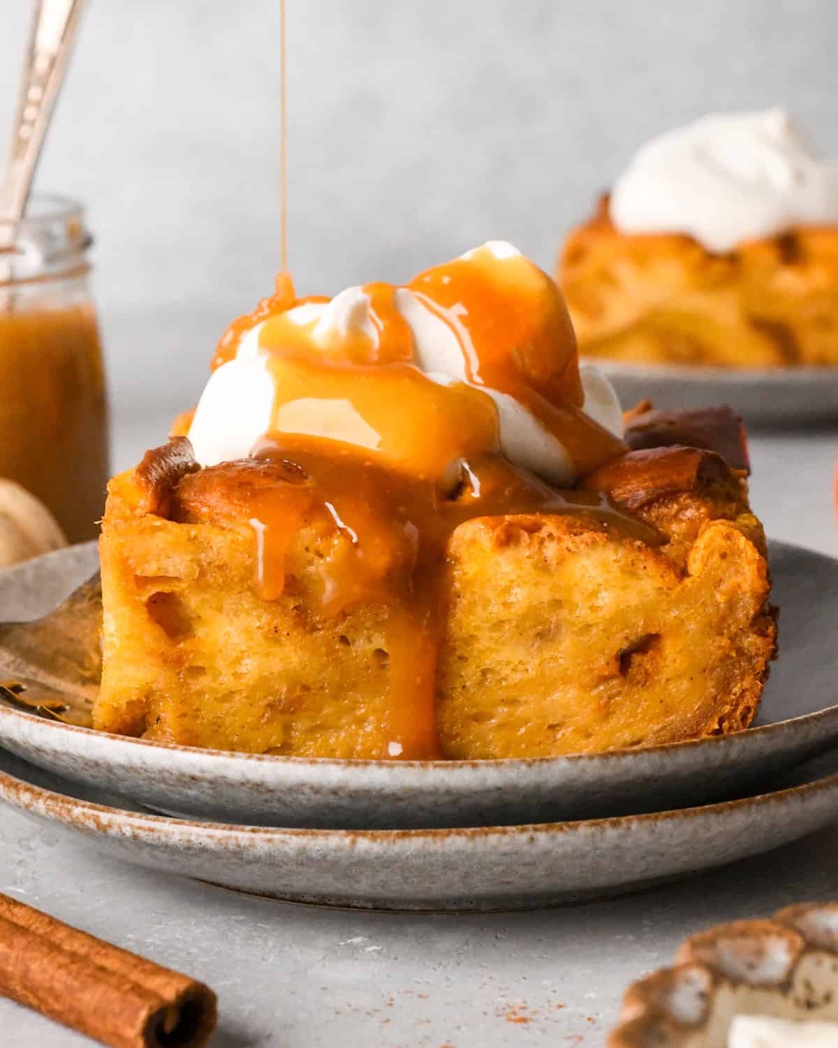 caramel sauce being drizzled over a piece of Pumpkin Bread Pudding on a plate topped with whipped cream. Another piece on a plate in the background, surrounded by cinnamon sticks and pumpkins