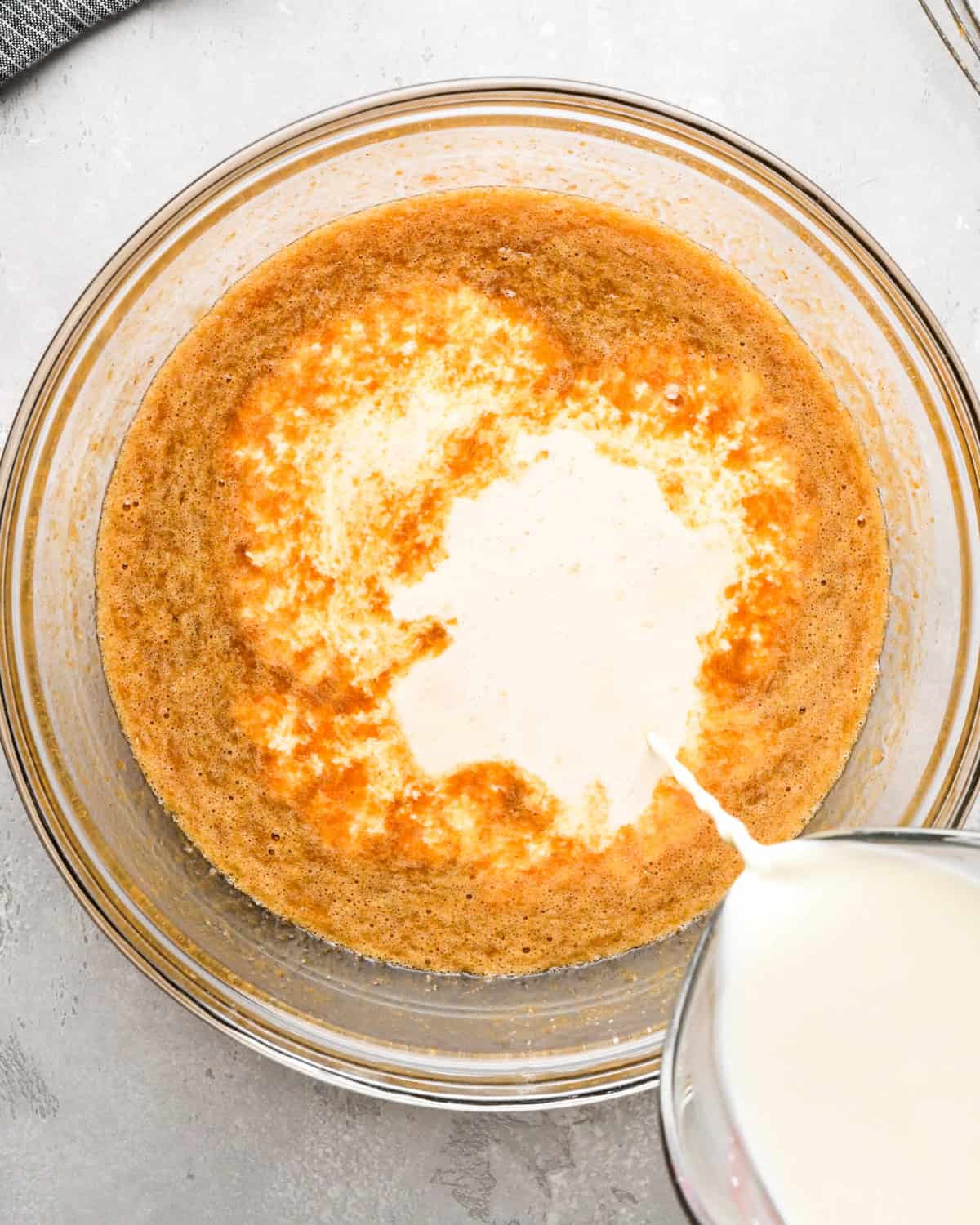 heavy cream being added to the pumpkin custard mixture in a glass bowl on a gray surface 