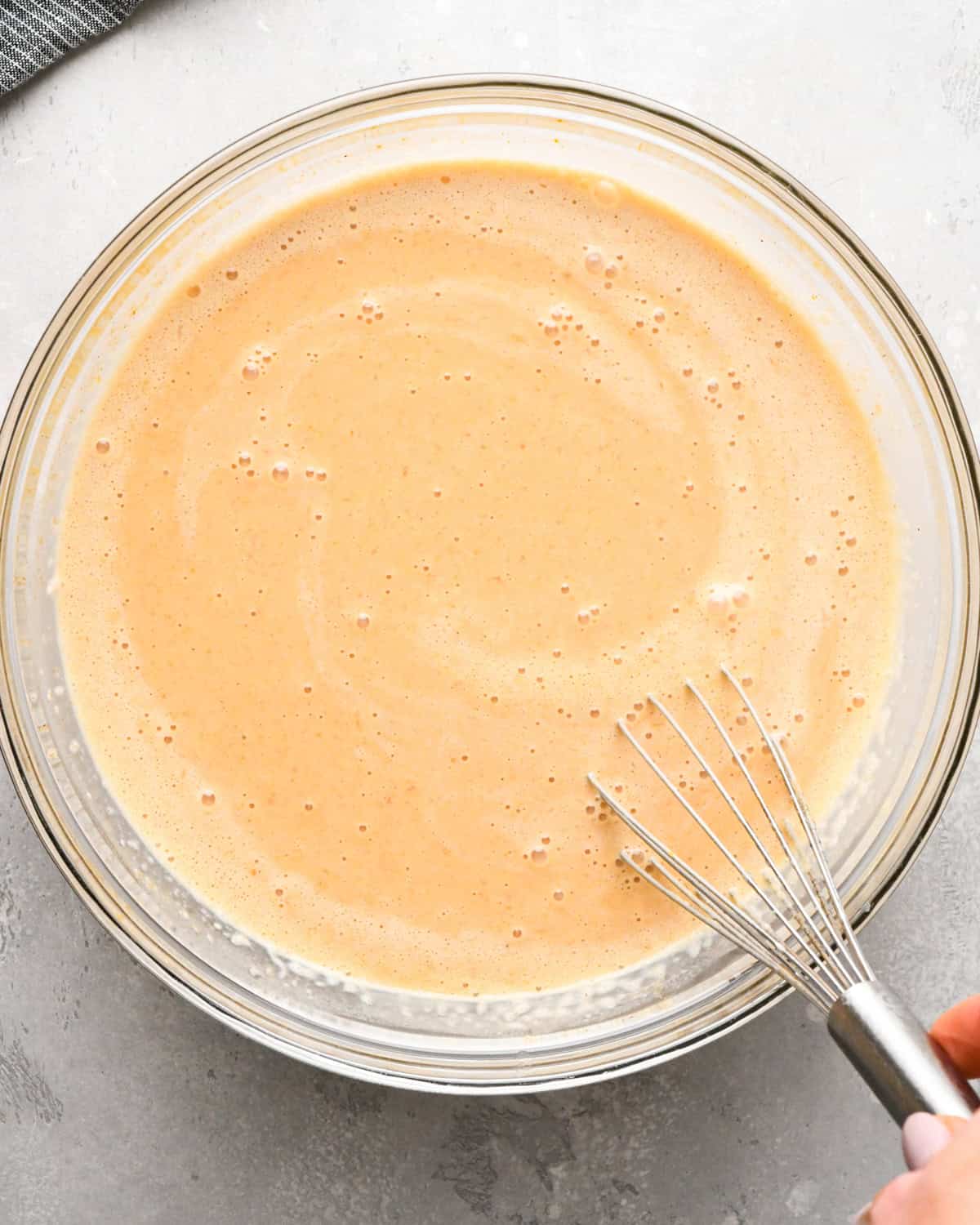 a whisk mixing the final pumpkin custard mixture in a glass bowl on a gray surface making pumpkin bread pudding
