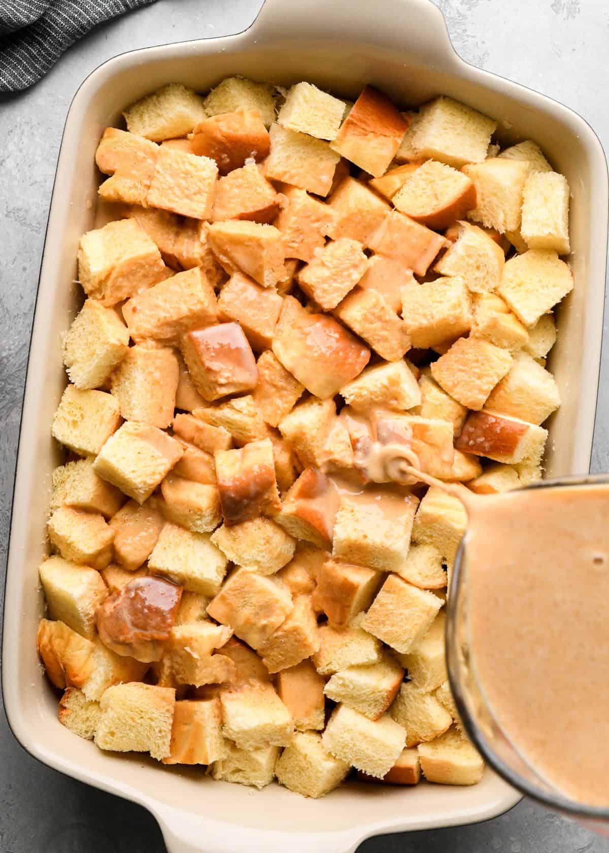 pumpkin custard being poured over cubed brioche bread in a 9x13" baking dish making pumpkin bread pudding