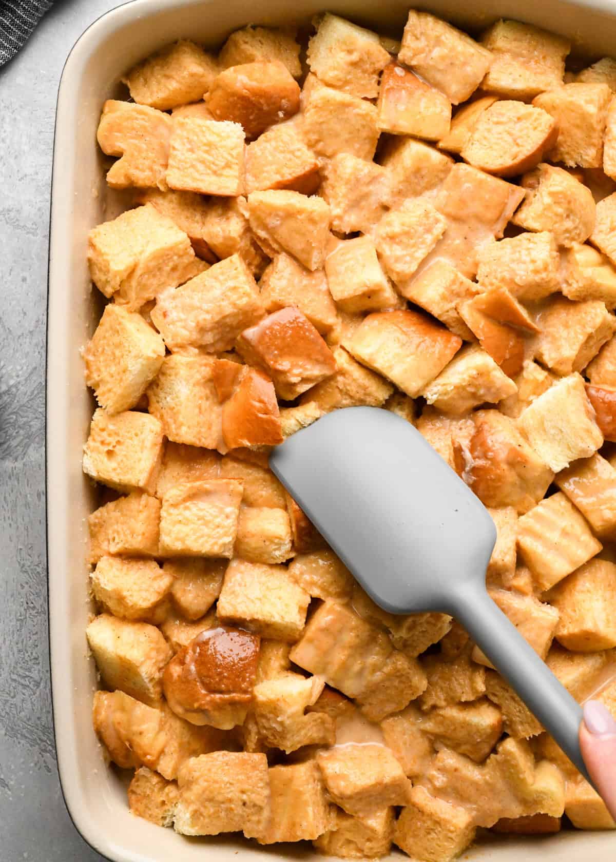 a spatula pressing down cubed brioche bread soaked in pumpkin custard in a baking dish on a gray surface making pumpkin bread pudding
