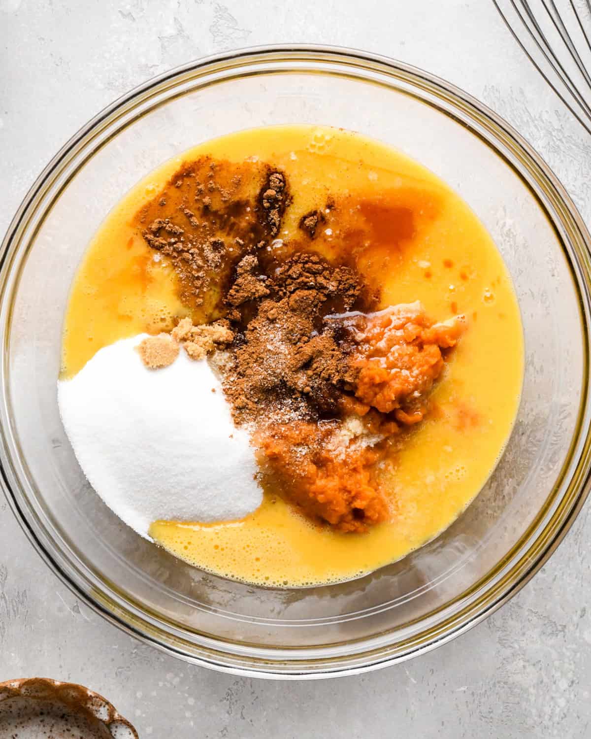 eggs, pumpkin puree, sugars, vanilla and cinnamon in a glass mixing bowl on a gray surface before mixing, making pumpkin bread pudding