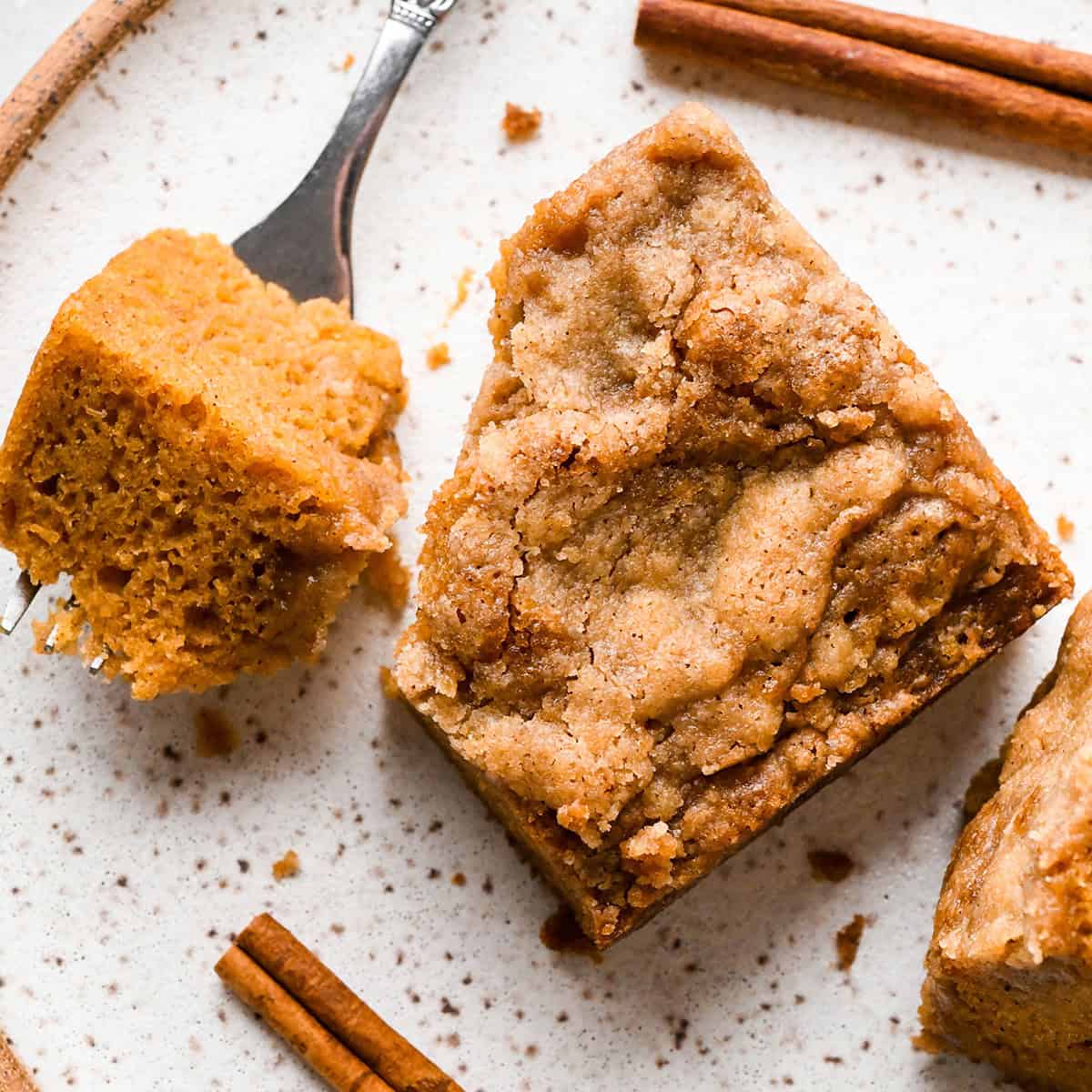 a piece of Pumpkin Coffee Cake on a white plate with a bite on a fork next to it styled with two cinnamon sticks