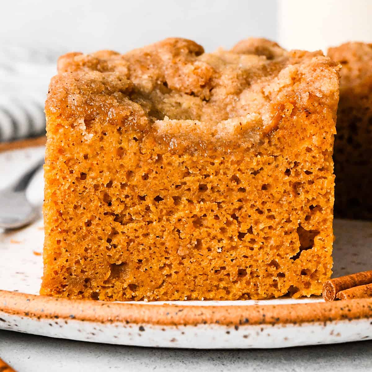 a piece of Pumpkin Coffee Cake on a white plate with a cinnamon stick and another piece of coffee cake int he background