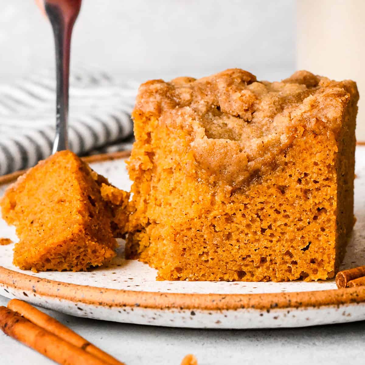 a piece of Pumpkin Coffee Cake on a white plate with a fork on the side with a bite on it styled with cinnamon sticks and a striped towel