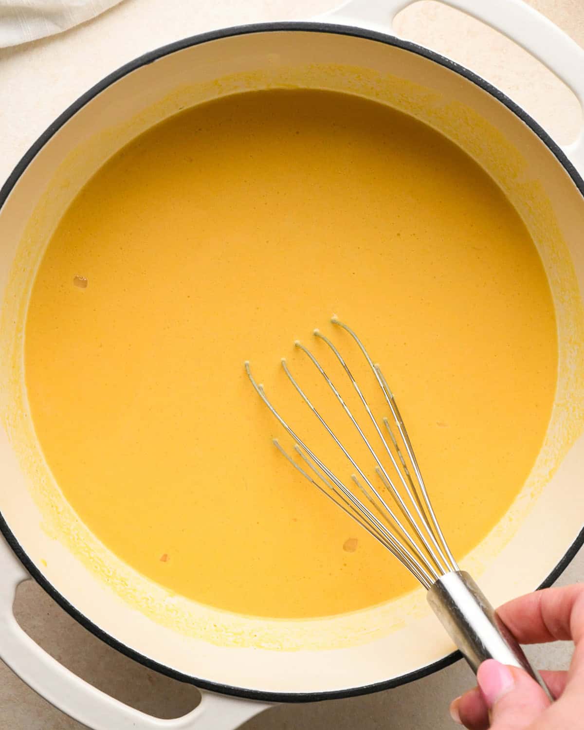 making pumpkin macaroni and cheese sauce in a white pan, a hand has whisked in the pumpkin with a silver whisk