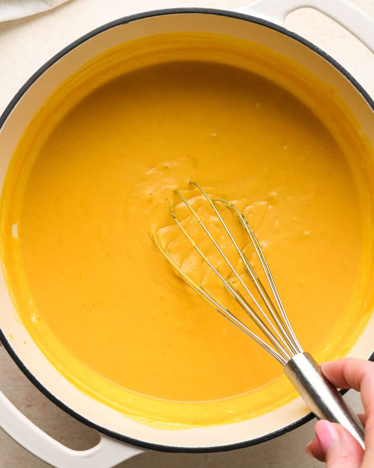 a hand holding a whisk in pumpkin mac and cheese sauce in a white pot to show that it has thickened. 