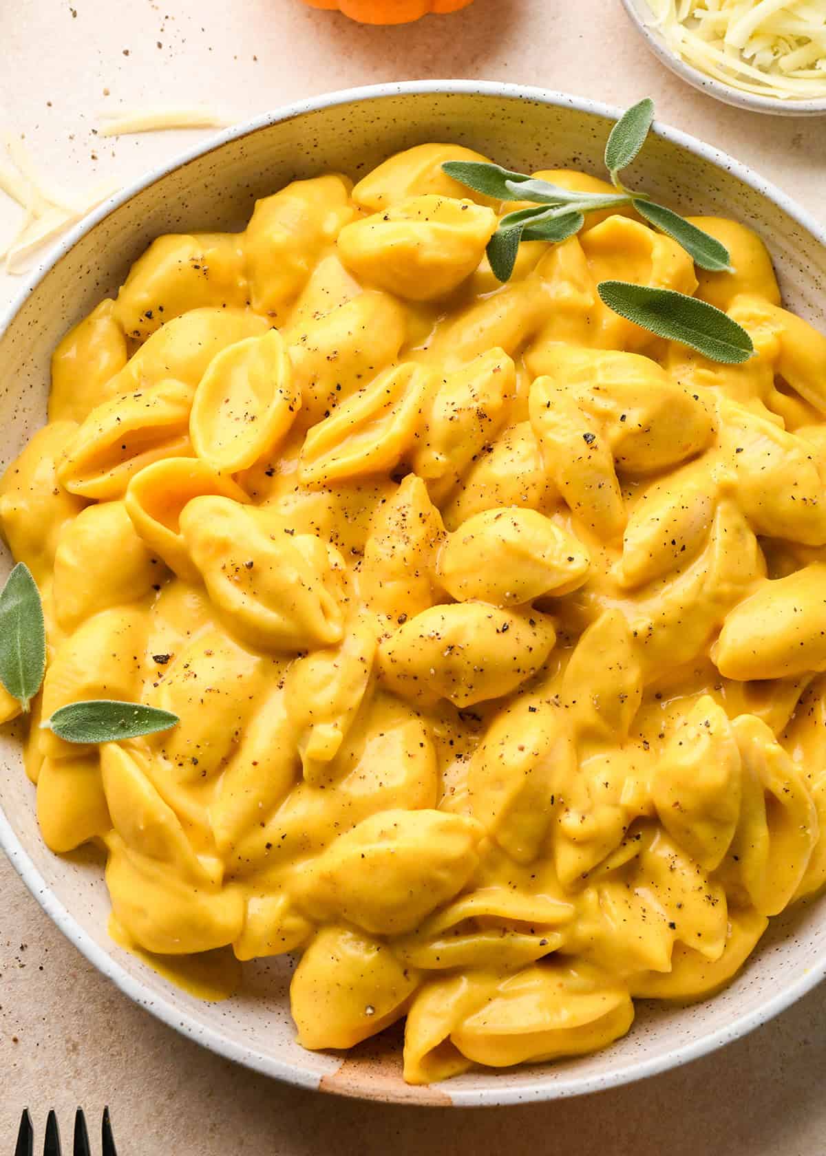 close-up of creamy pumpkin macaroni and cheese made with shell pasta sprinkled with black pepper and garnished with fresh sage leaves in a speckled ceramic bowl, with shredded cheese, a fork and a mini pumpkin in the background.