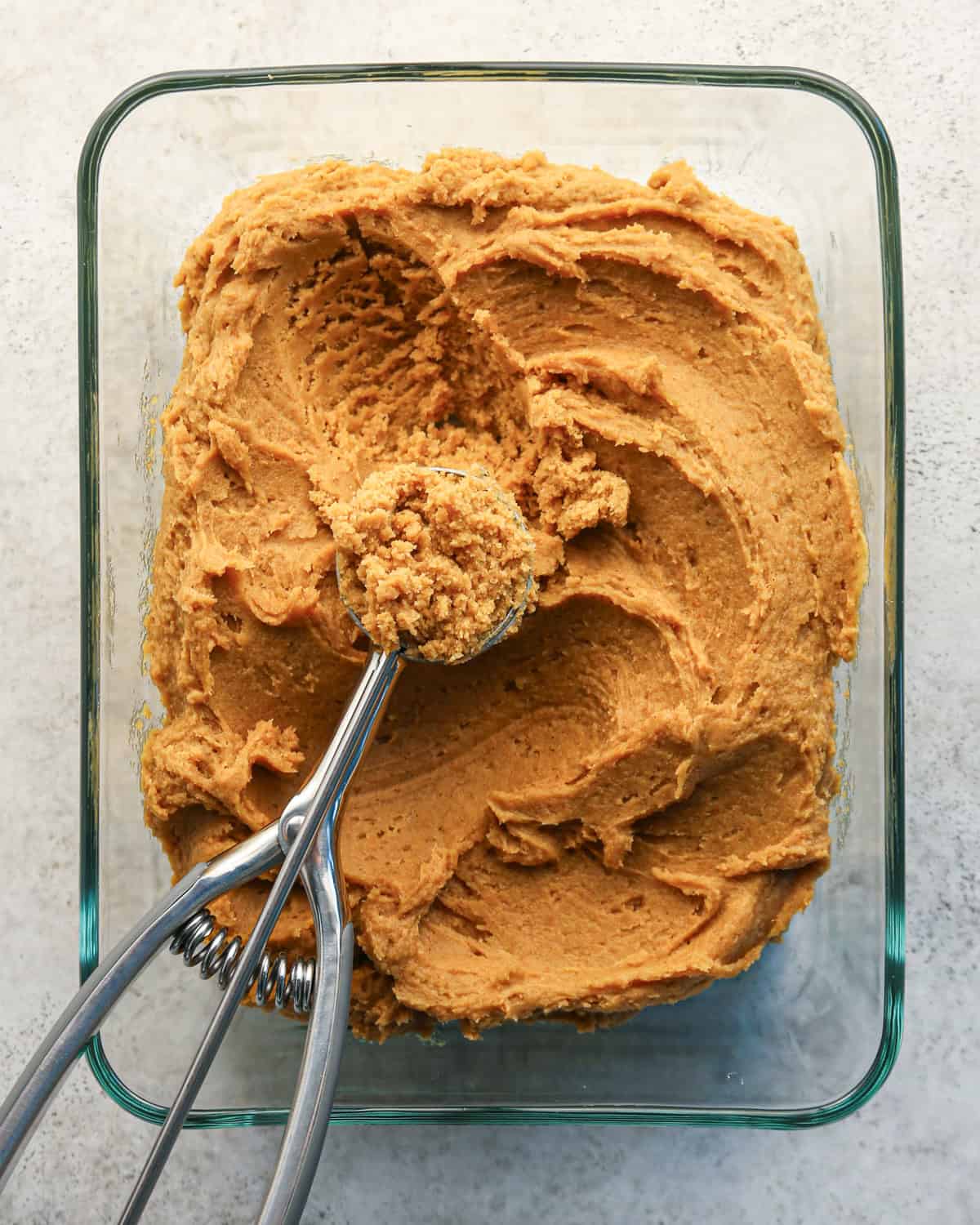 pumpkin snickerdoodle cookie dough after chilling in a rectangular glass container on a gray surface with a cookie scoop that has a scoop of dough in it on a gray surface