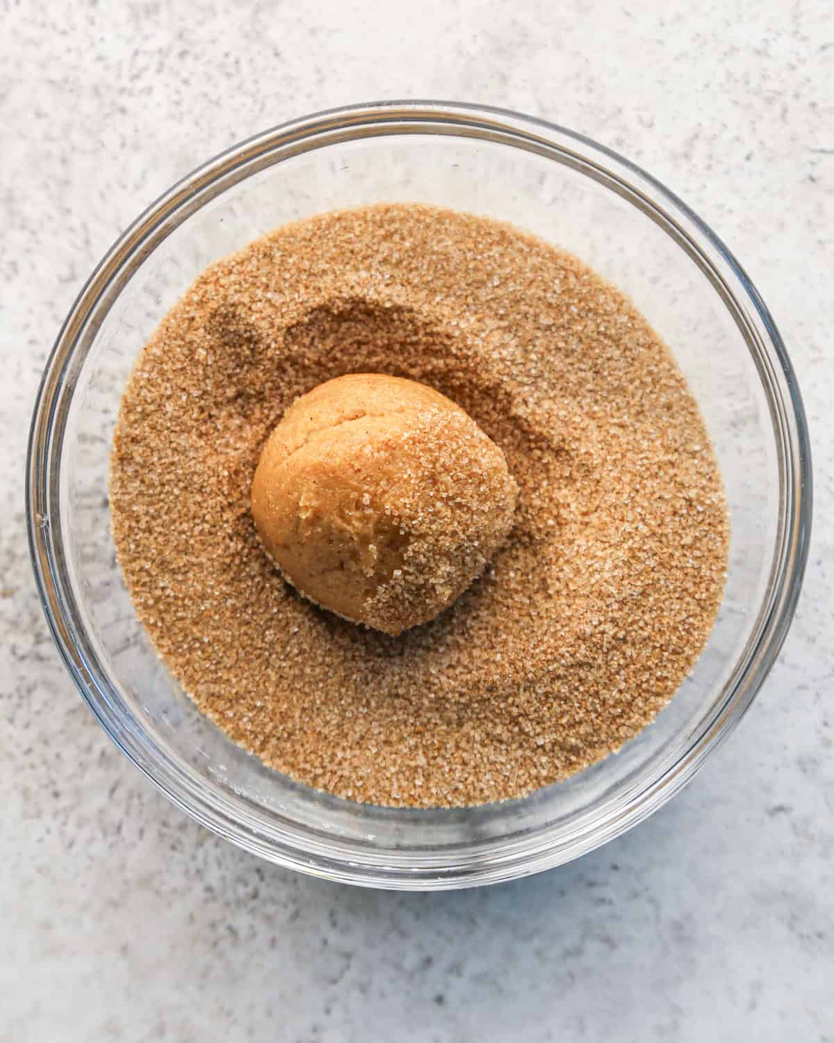 a ball of pumpkin snickerdoodle cookie dough in a glass bowl of cinnamon sugar on a gray surface
