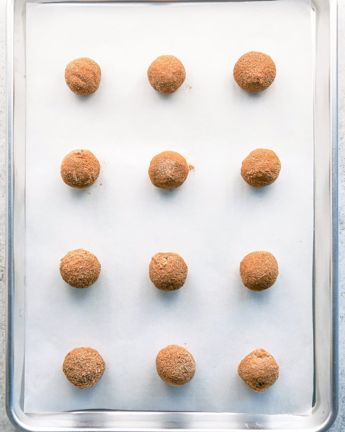 12 pumpkin snickerdoodle cookie dough balls covered in cinnamon sugar on a silver baking sheet lined with parchment paper before baking