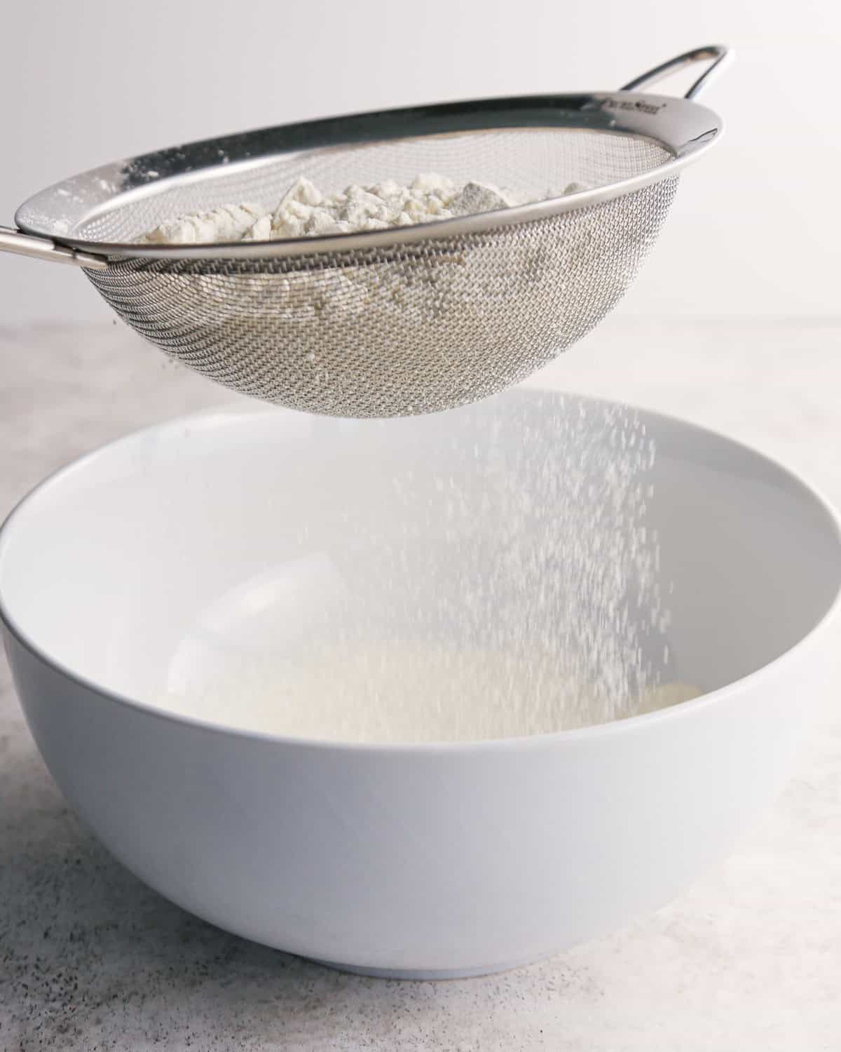 flour being sifted in a fine mesh metal strainer over a white bowl to make pumpkin snickerdoodles
