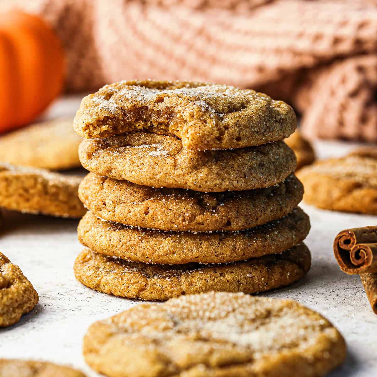 a stack of 5 of pumpkin snickerdoodle cookies coated in cinnamon sugar, with a bite taken out of the top cookie. Surrounded by a pumpkin, cinnamon sticks, a mauve towel and more cookies. 