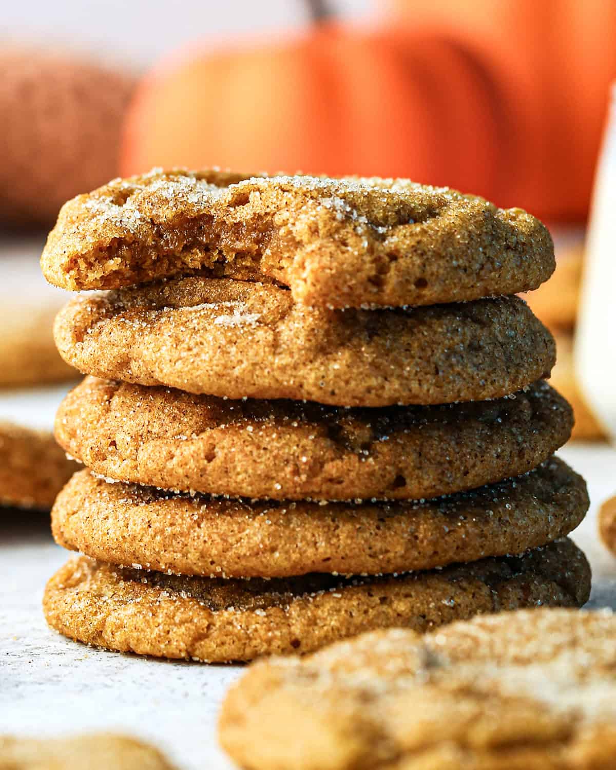 a stack of 5 of pumpkin snickerdoodle cookies coated in cinnamon sugar, with a bite taken out of the top cookie. Blurred pumpkins, more cookies and a glass of milk in the background.