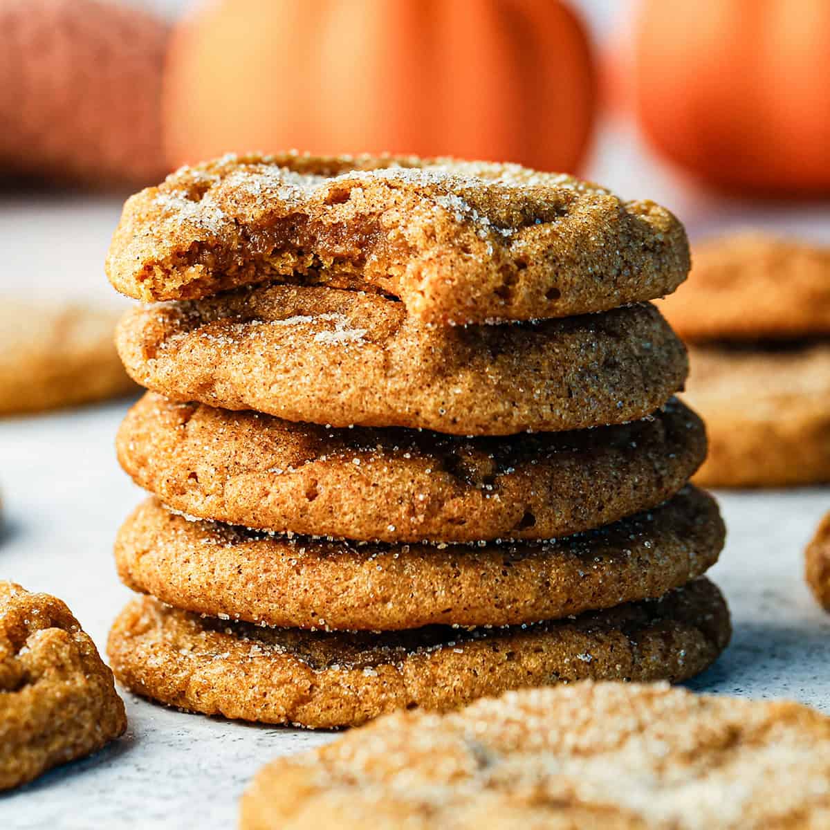 a stack of 5 of pumpkin snickerdoodle cookies coated in cinnamon sugar, with a bite taken out of the top cookie. Blurred pumpkins, more cookies and a glass of milk in the background.
