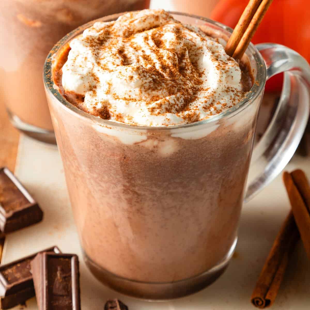 A glass mug filled with pumpkin hot chocolate, topped with a swirl of whipped cream, dusted with pumpkin spice, and garnished with a cinnamon stick. Surrounded by dark chocolate chunks and whole cinnamon sticks with a pumpkin in the background