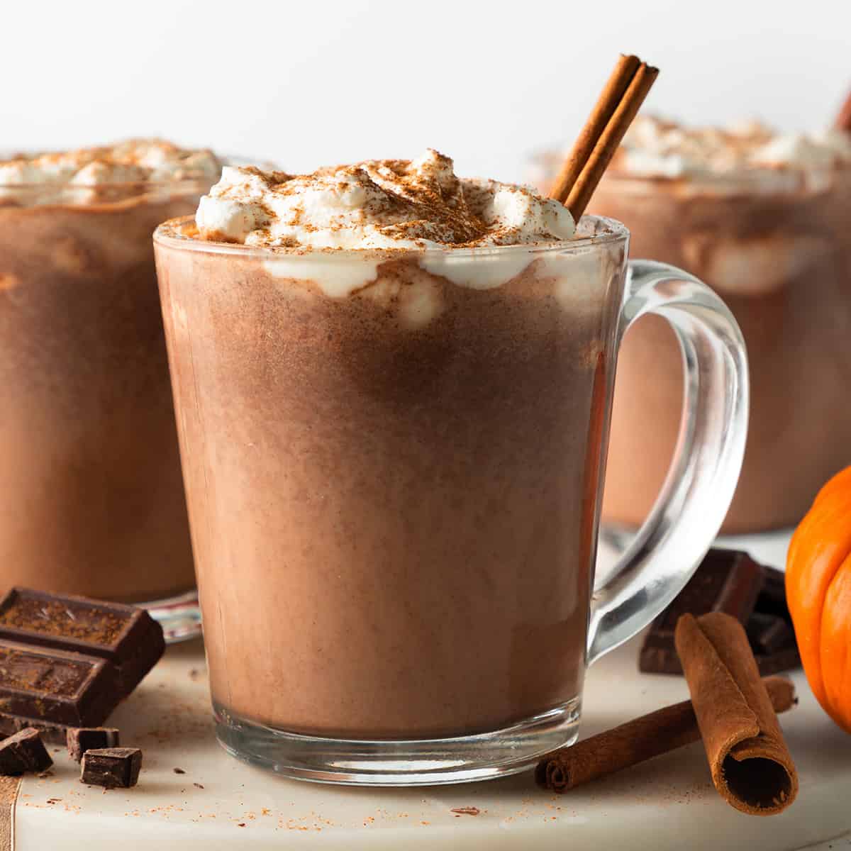 3 glass mugs filled with pumpkin hot chocolate, topped with a swirl of whipped cream, dusted with pumpkin spice, and garnished with a cinnamon stick. Surrounded by dark chocolate chunks and whole cinnamon sticks with a pumpkin in the background