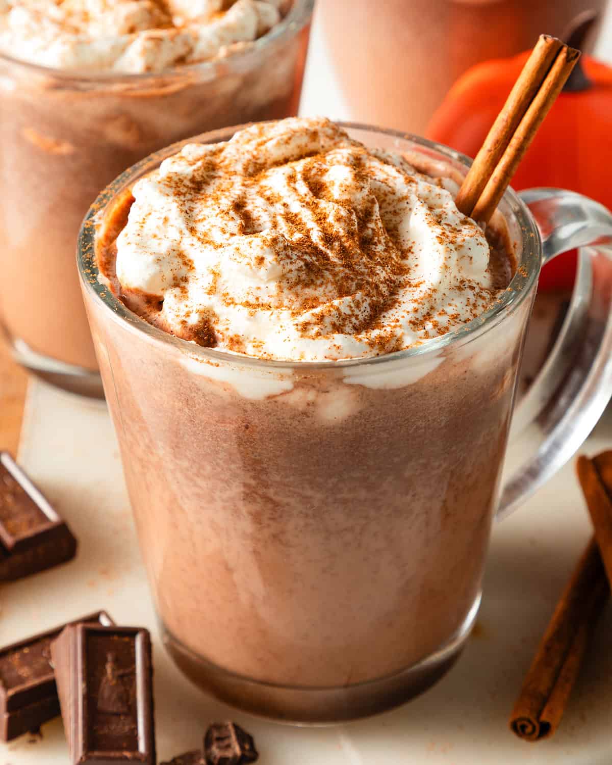 2 glass mugs filled with pumpkin hot chocolate, topped with a swirl of whipped cream, dusted with pumpkin spice, and garnished with a cinnamon stick. Surrounded by dark chocolate chunks and whole cinnamon sticks with a pumpkin in the background