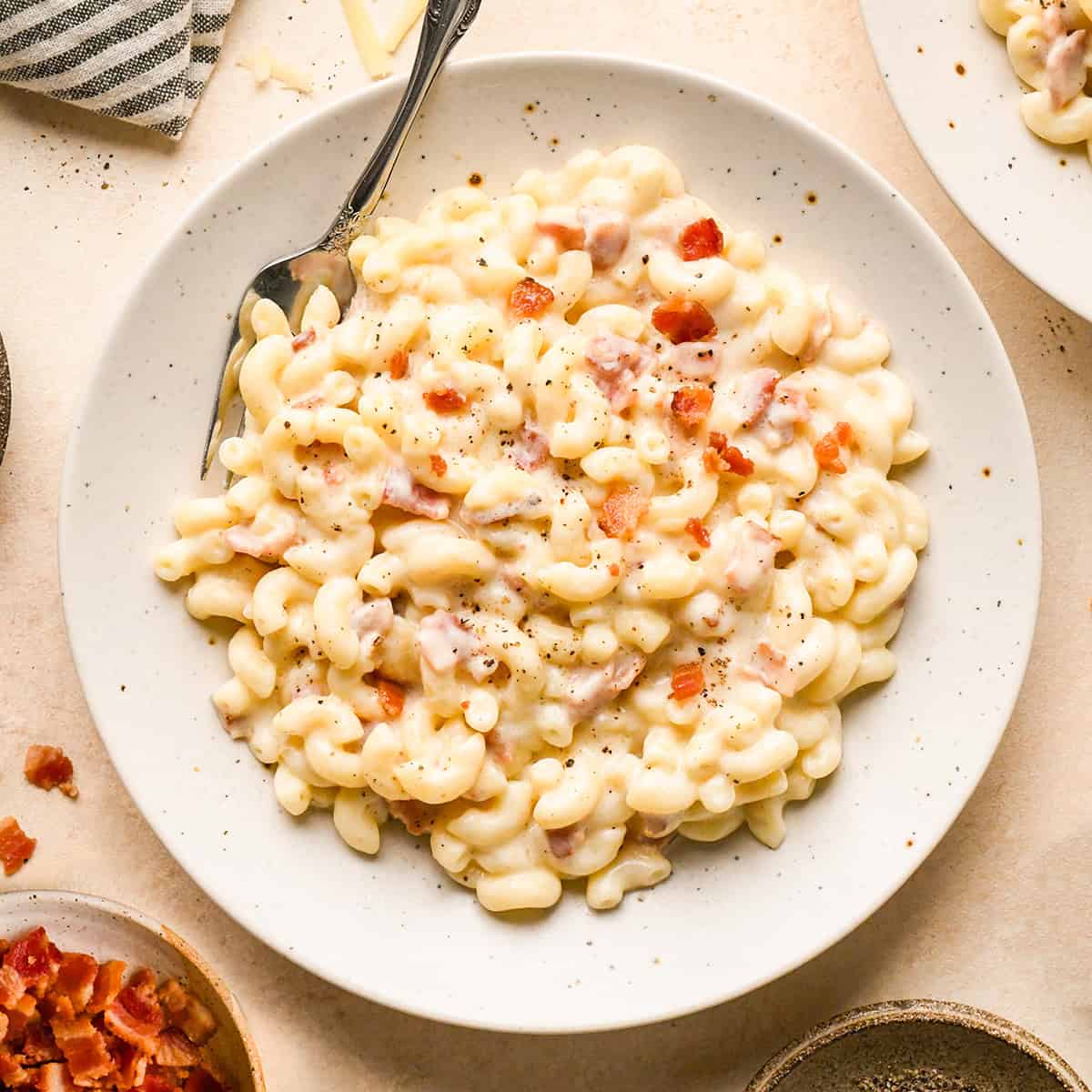 Bacon macaroni and cheese on a white speckled plate, topped with cooked bacon pieces and freshly ground black pepper, with a fork resting on the side.
